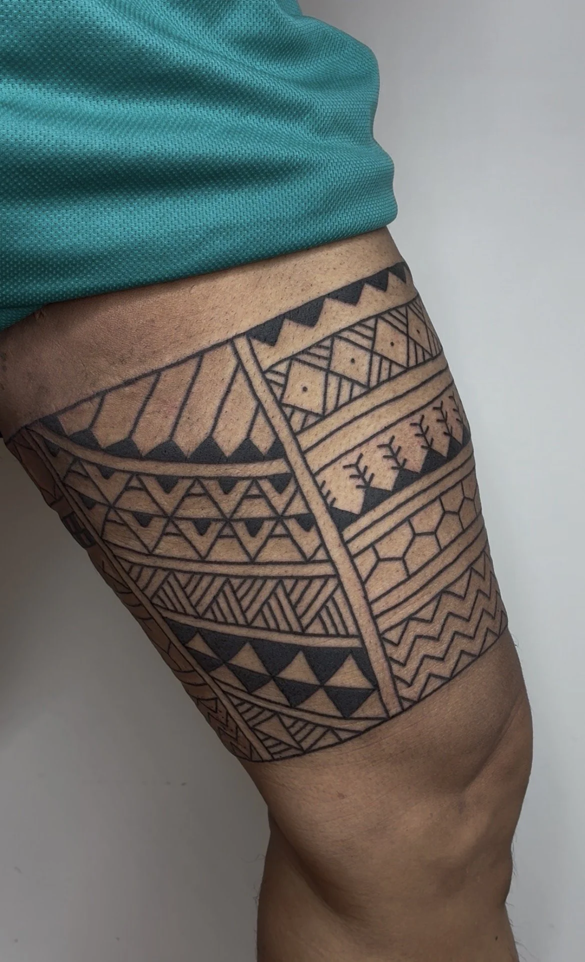 Close-up of a person's upper thigh with traditional black tribal tattoo patterns, wearing a teal athletic shirt.