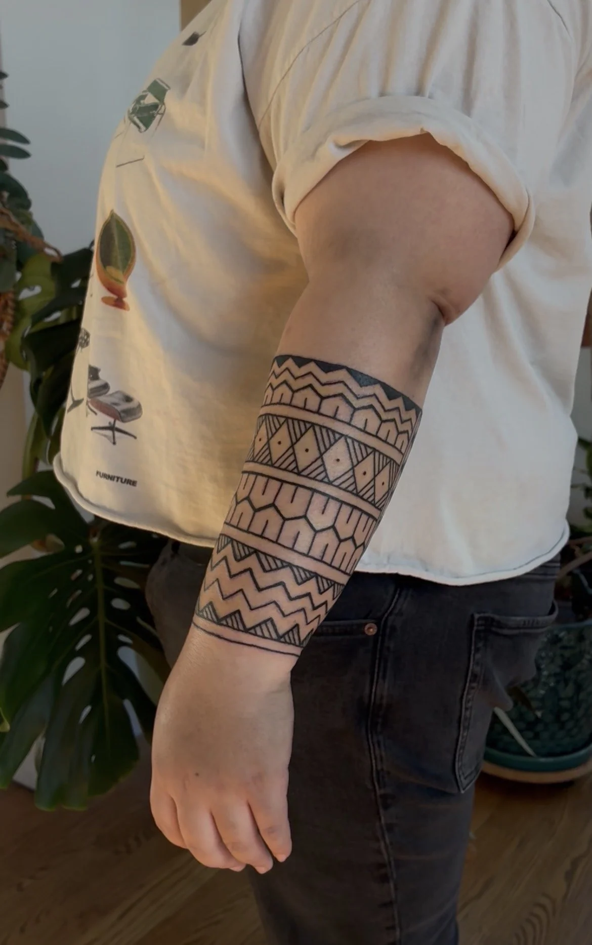 Person wearing a white t-shirt with colorful graphic prints on the sleeve and a black geometric tattoo on their arm, standing near a plant with large green leaves.