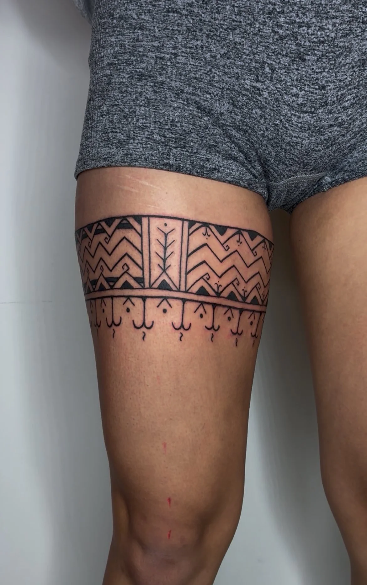 Close-up of a person's upper thigh with a tattoo featuring geometric patterns and symbols, wearing gray shorts.
