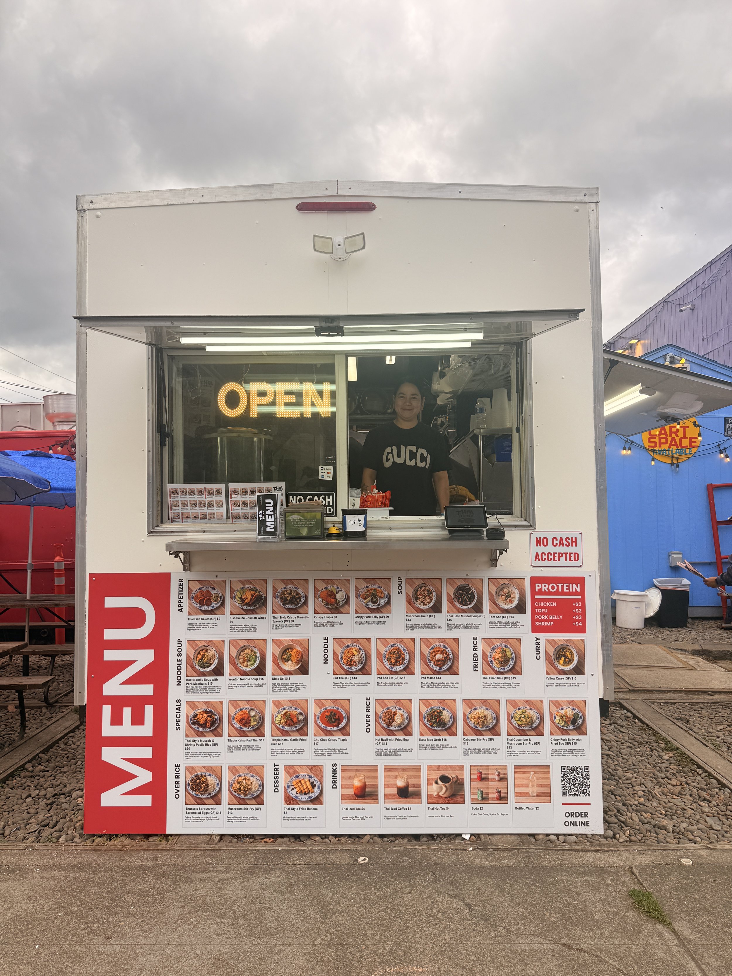 Front view of the Thai Station PDX food cart in Sellwood, Portland, featuring a Thai street food menu, colorful food photos, an open service window, and a welcoming staff member inside.