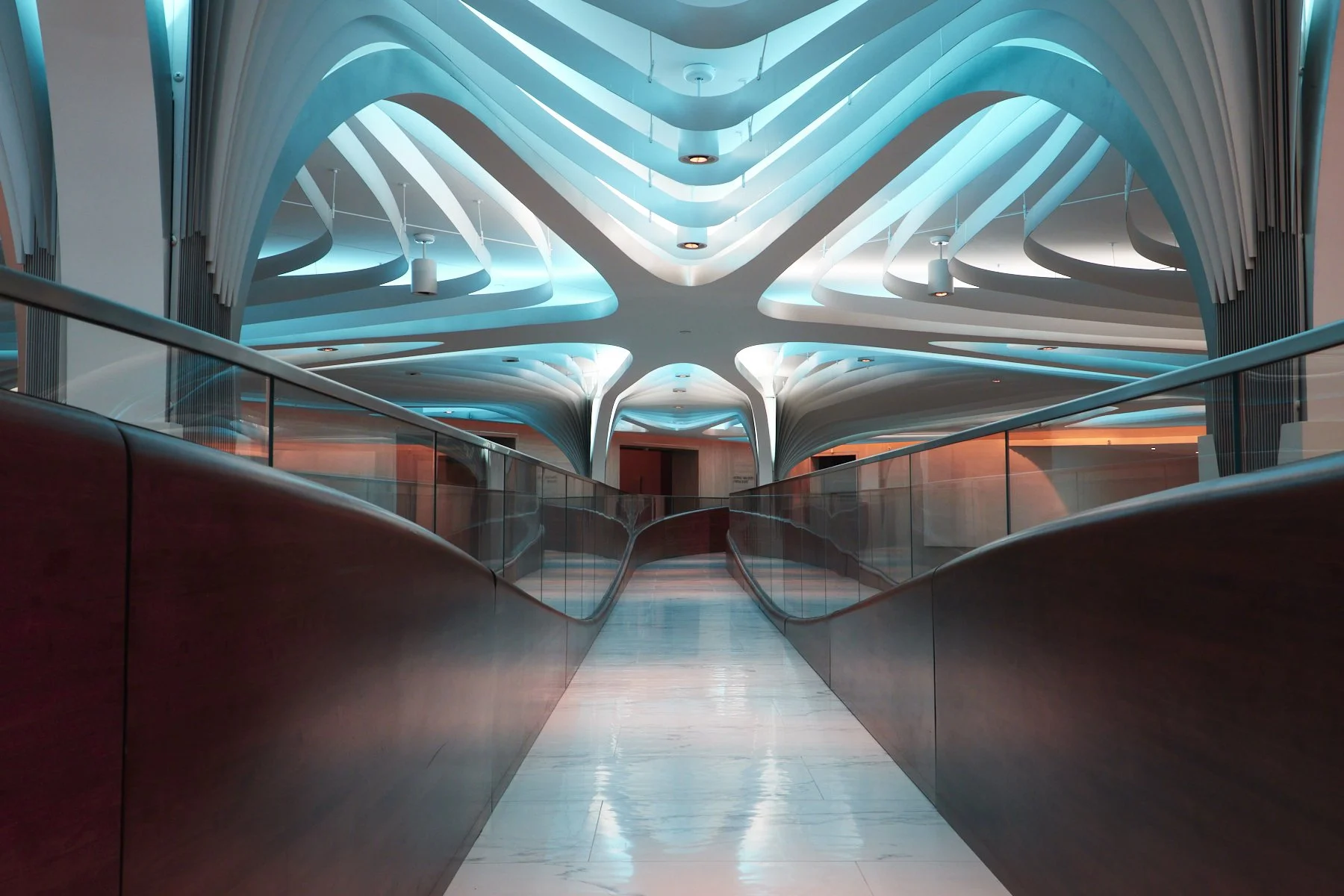 Modern interior with futuristic curved ceiling structures illuminated in blue and white, viewed from a walking path with glass railings.