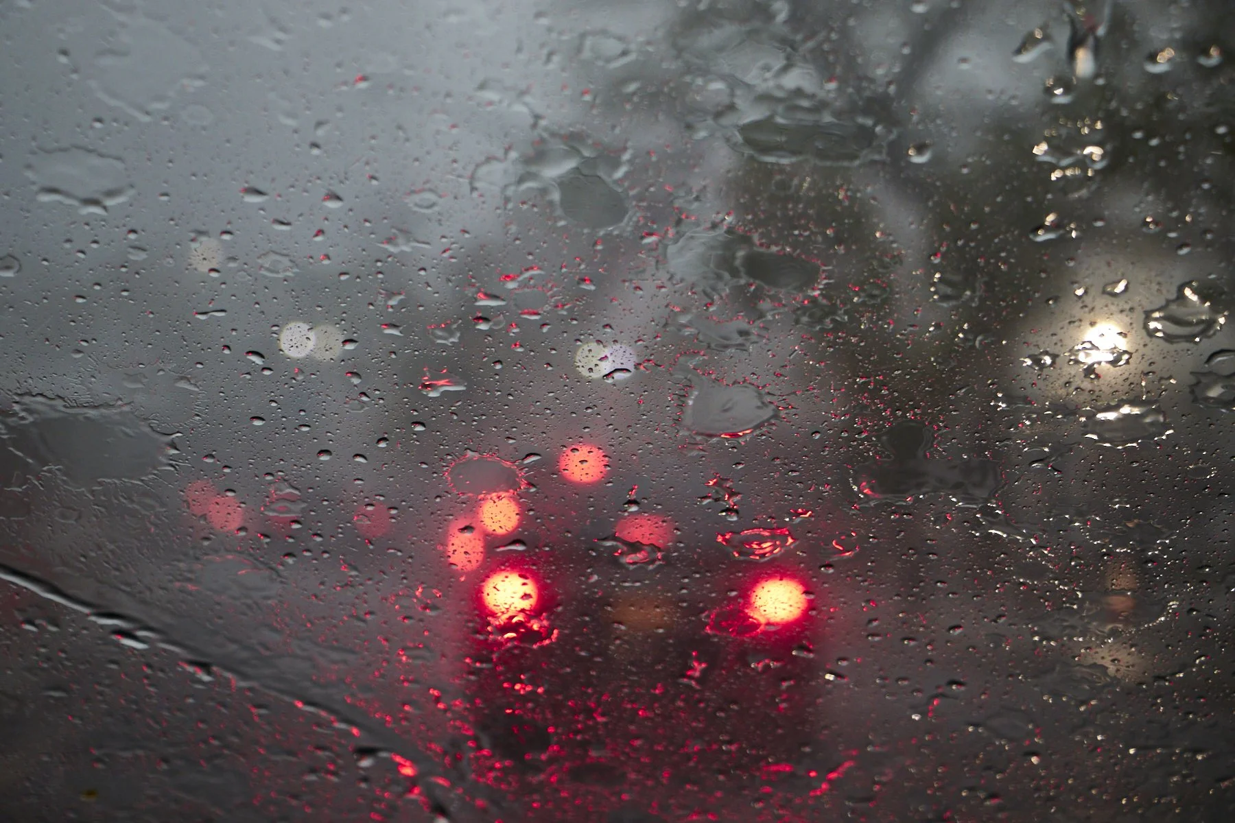 Rain-covered car windshield with blurred red and yellow tail lights of other vehicles in traffic on a cloudy day.