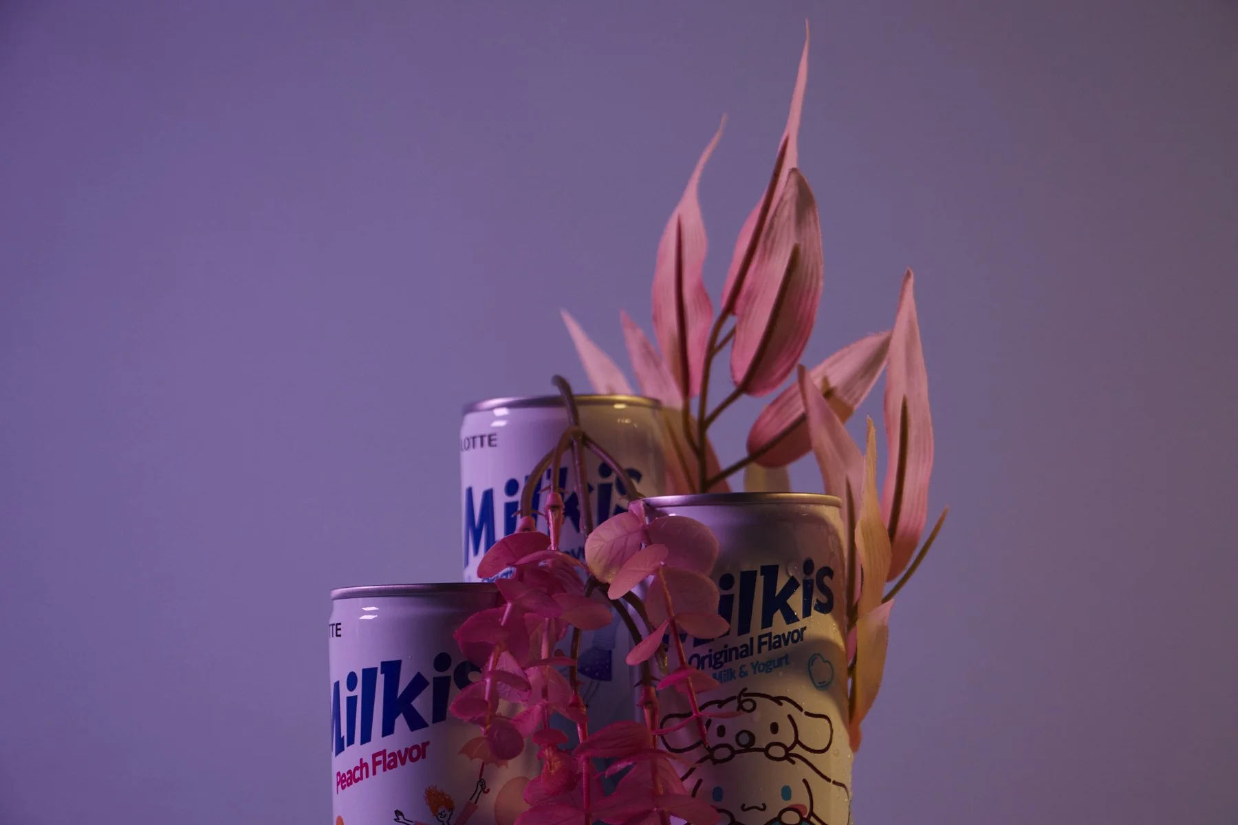 Three cans of Milkis Peach Flavor soda with pink and yellow flowers arranged above and around them, set against a purple background.