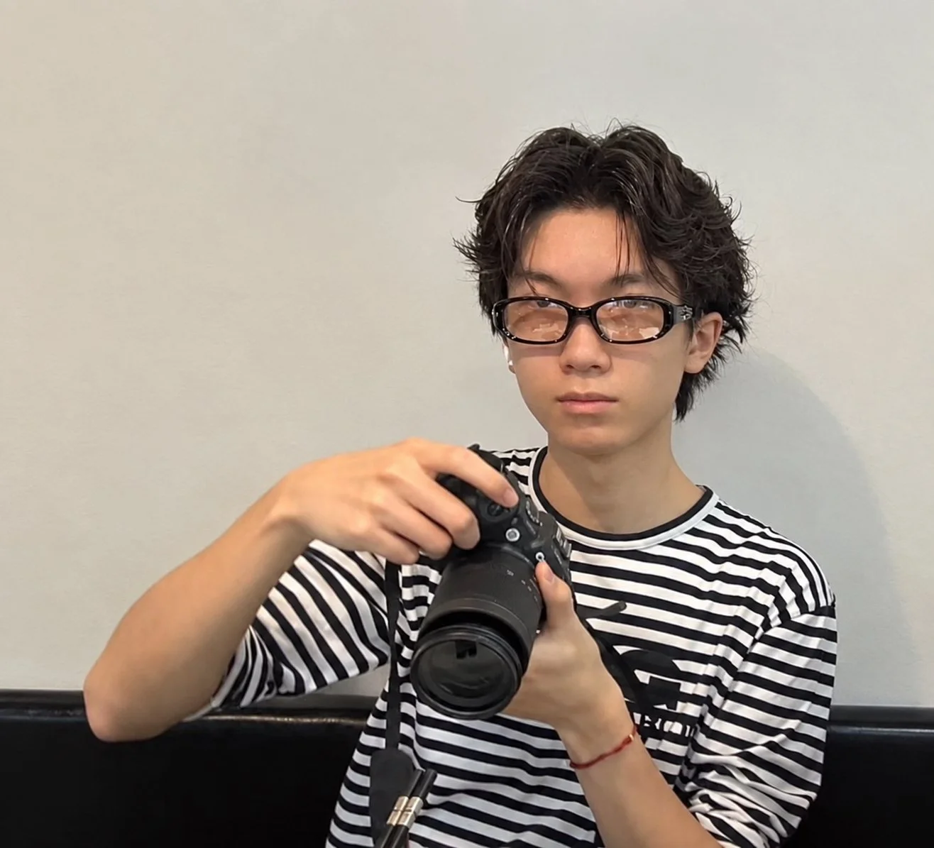 A young person with curly dark hair and glasses holding a video camera, wearing a black and white striped shirt, sitting against a plain white wall.