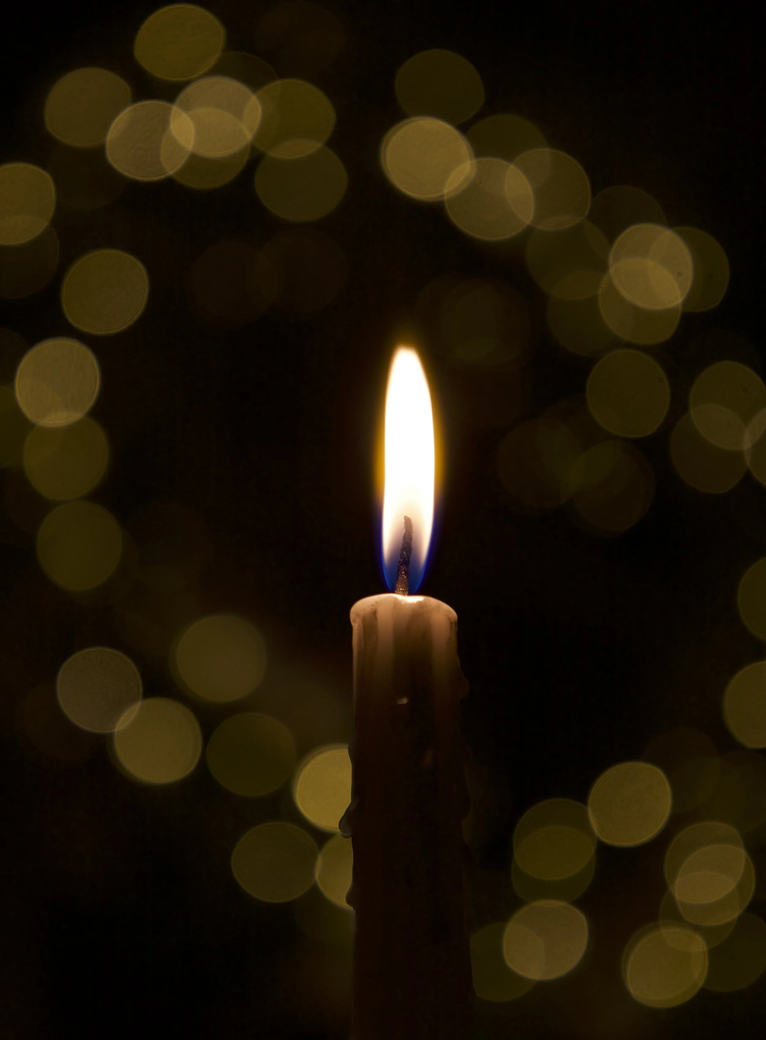 Image of a lit candle with a softly glowing flame, against a background of blurred golden bokeh lights.