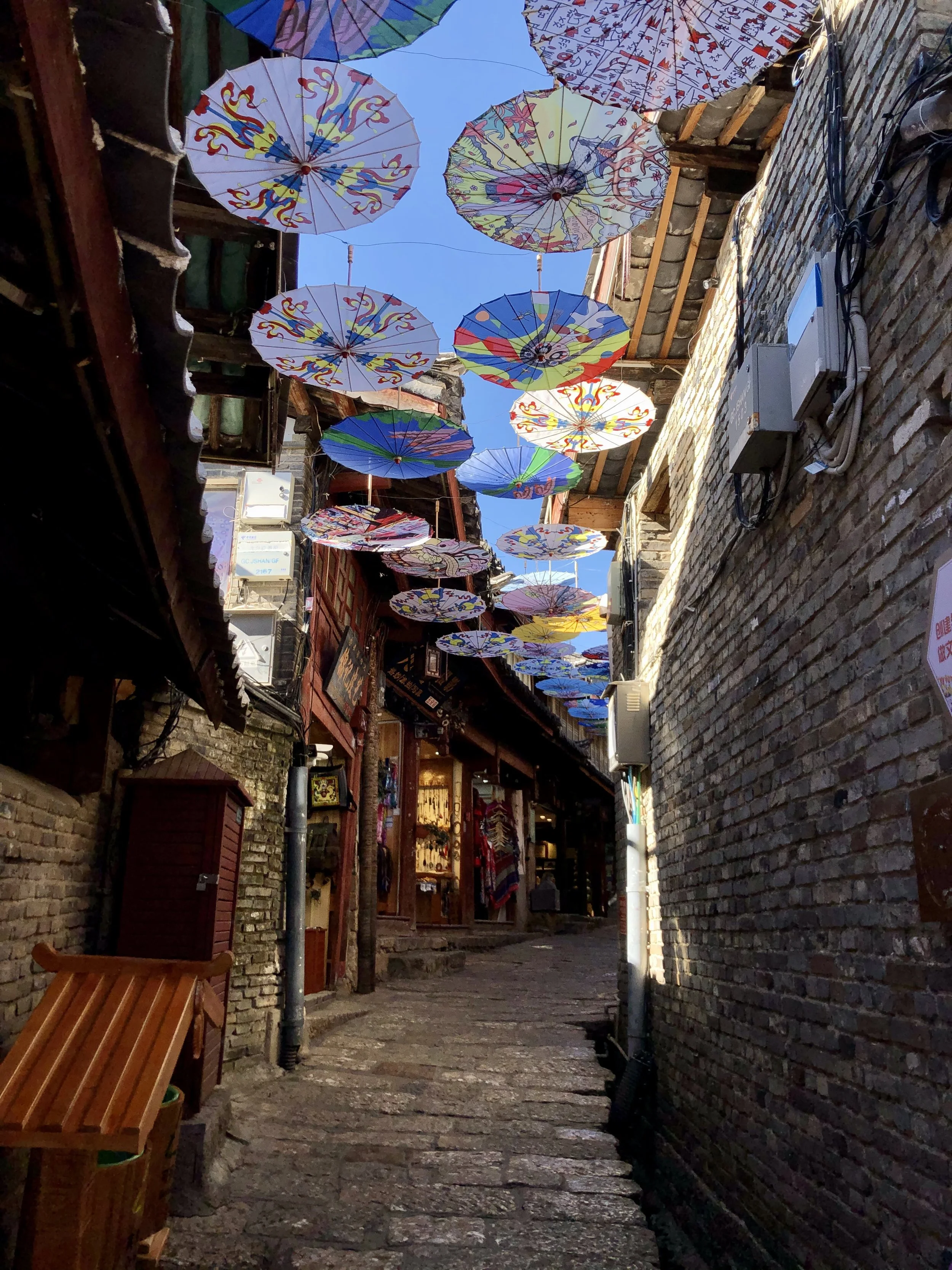 Narrow alleyway with brick walls on both sides, decorated with colorful paper umbrellas hanging overhead against a bright blue sky, with shops and wooden benches lining the alley.