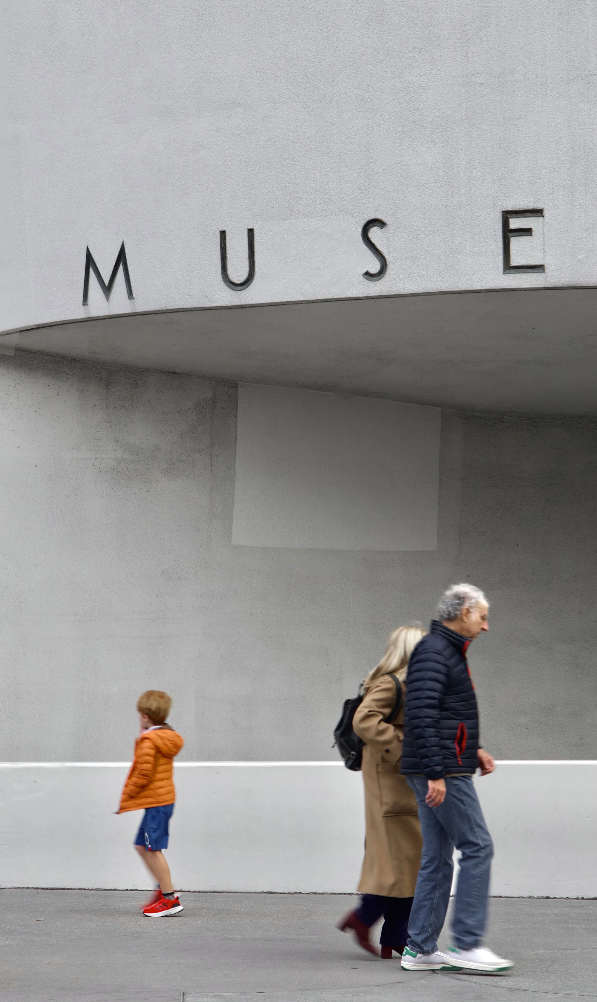 People walking past a modern building with the word 'MUSE' on it. The scene includes a child in an orange jacket, an older man in a navy jacket, and a woman in a tan coat.