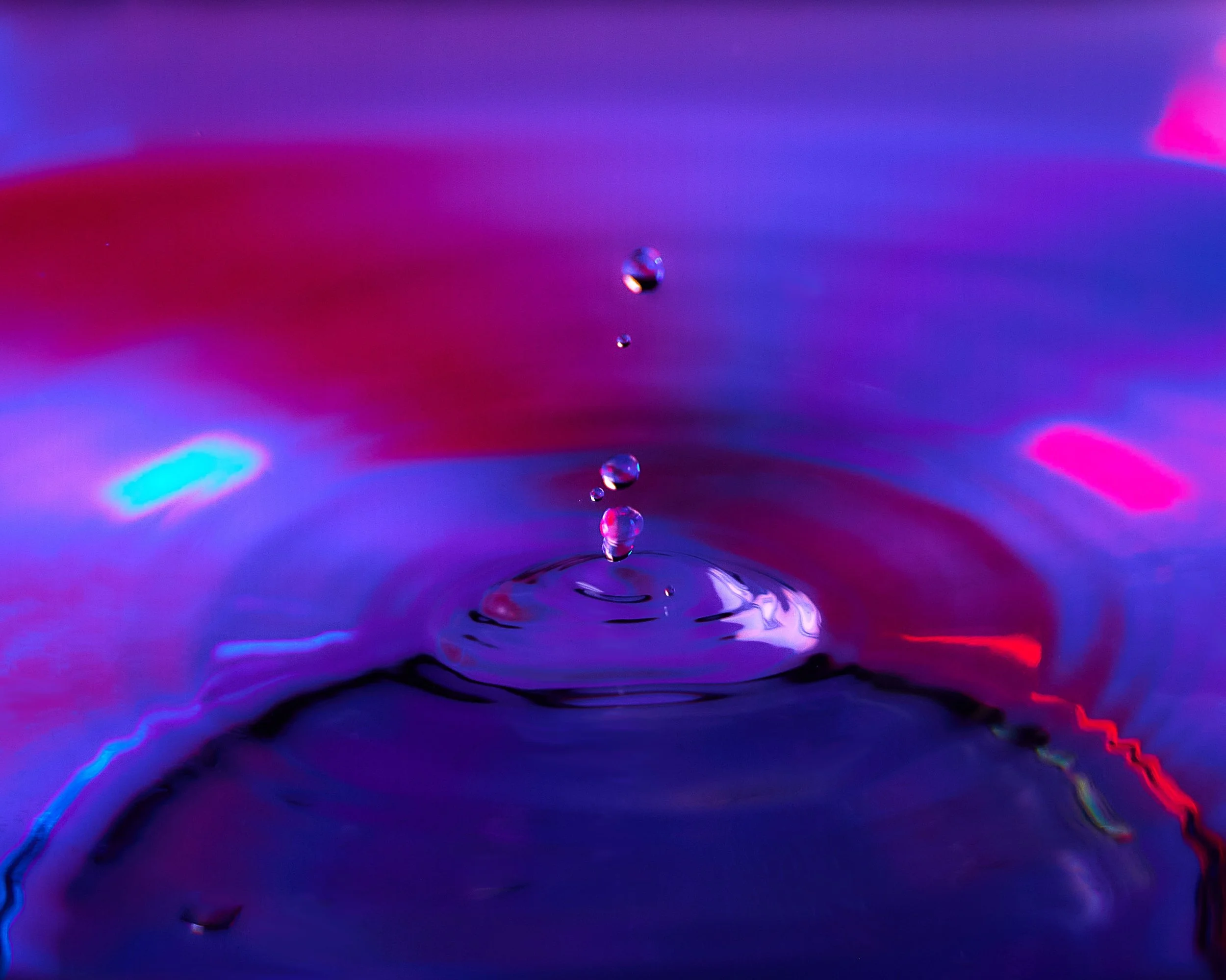 Close-up of water droplets falling into a liquid, creating ripples, with vibrant pink, purple, and blue background colors.