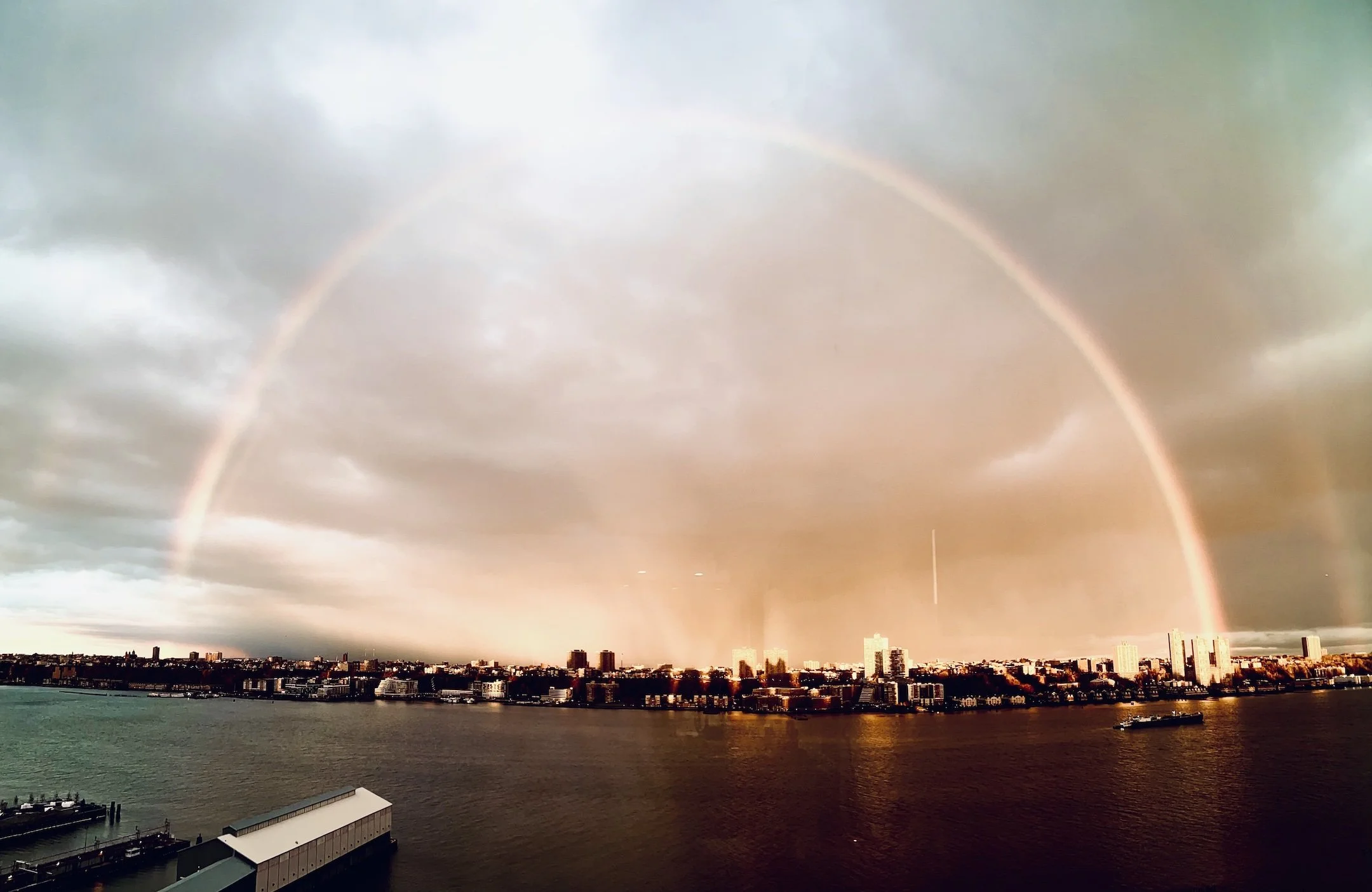 Multiple rainbows arch across a cloudy sky over a city skyline by a body of water at sunset.