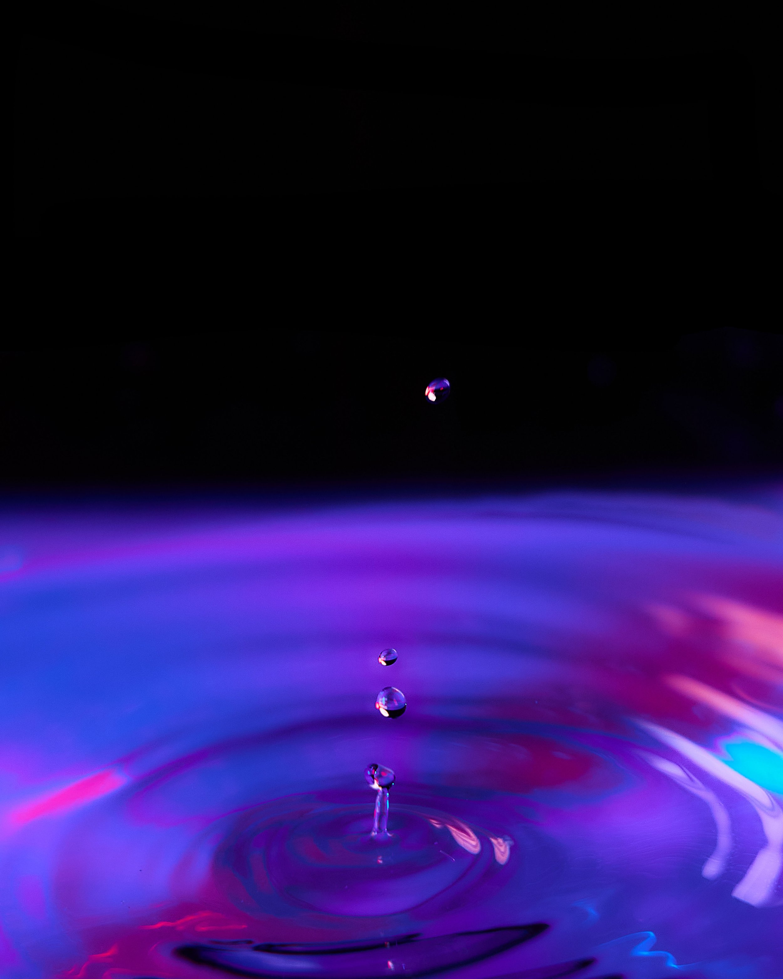 Close-up of a water droplet falling into a colorful liquid, creating ripples and a splash, with vibrant purple, blue, pink, and red hues.