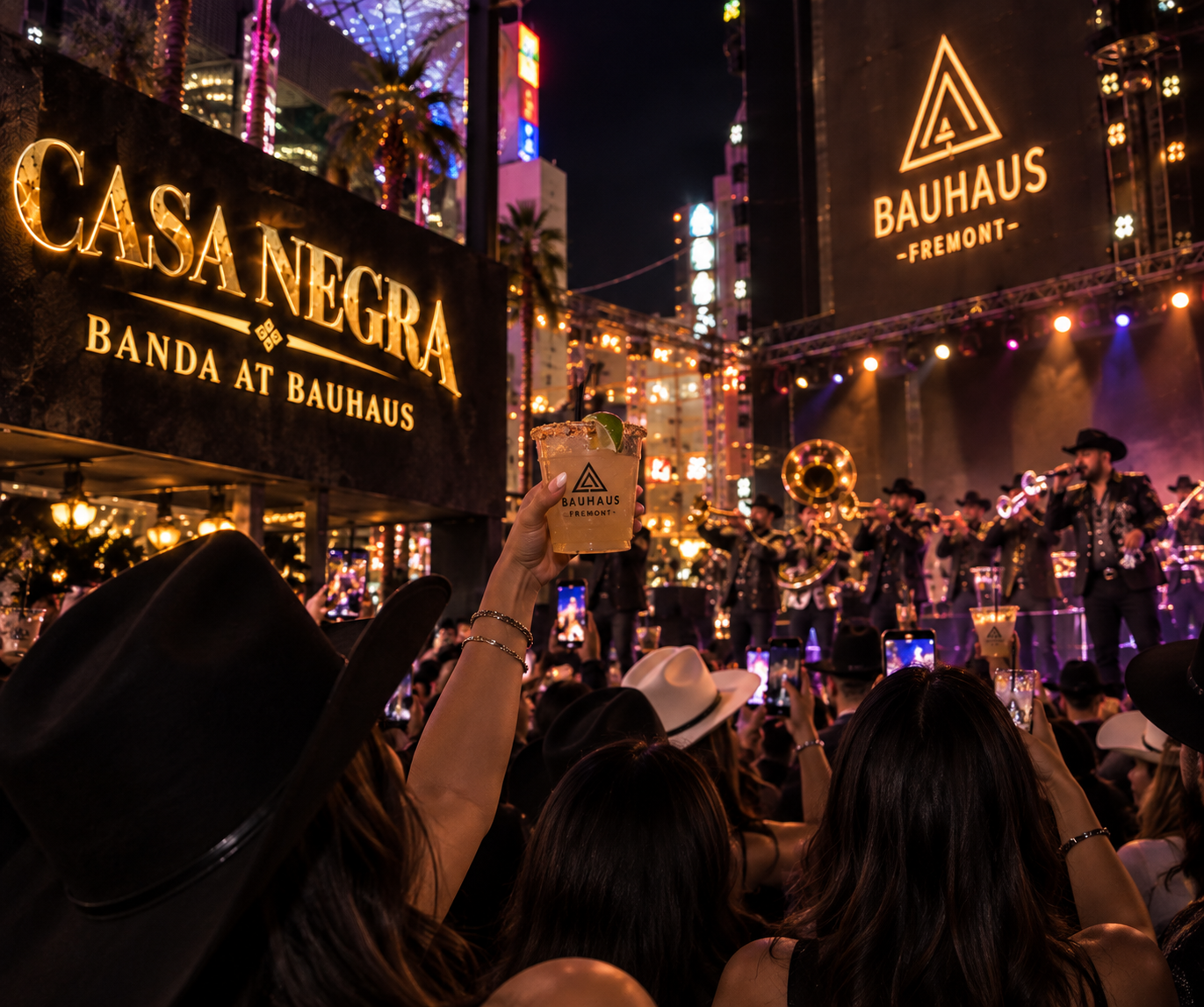 Crowd watching a live music performance at night, holding drinks, with bright neon signs and stage lights. One person in the foreground is holding a drink with a lime wedge.