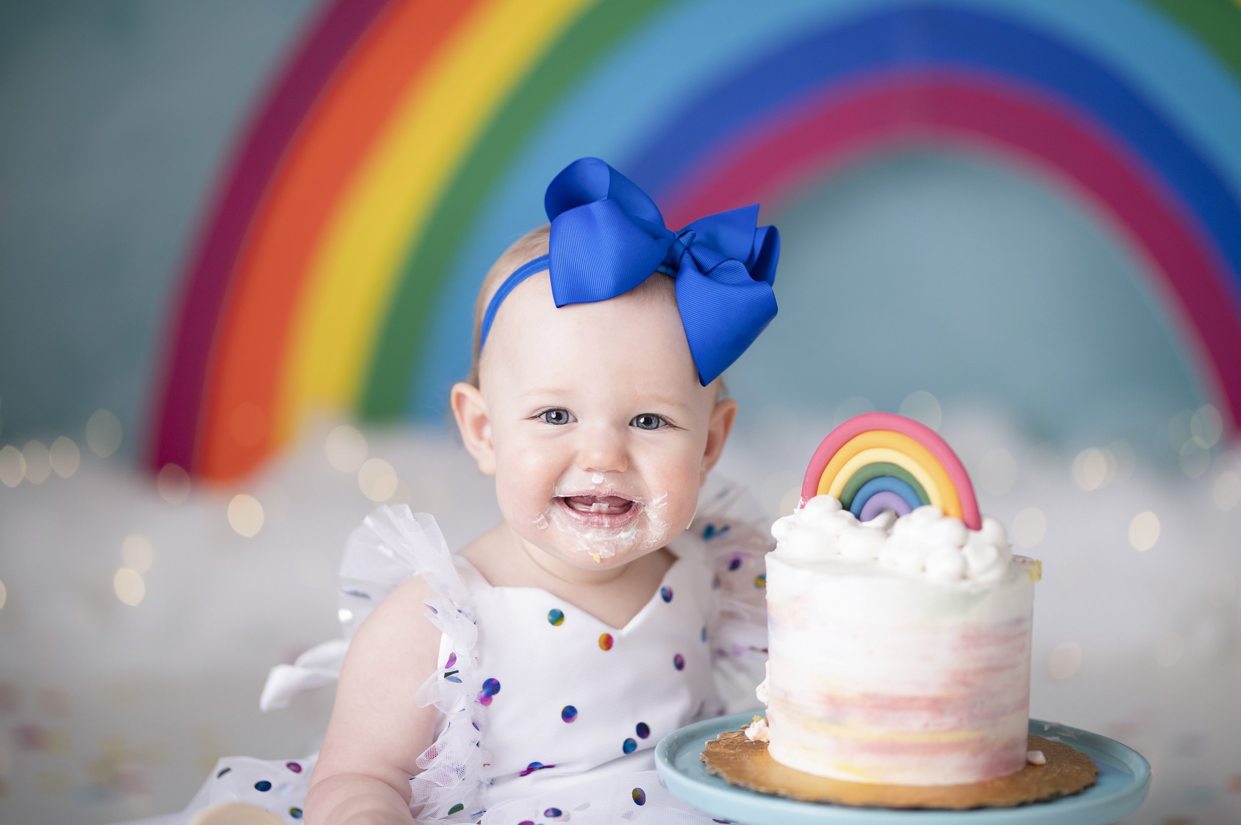 Smiling baby with a blue bow posing beside a small rainbow cake during a colorful birthday session.