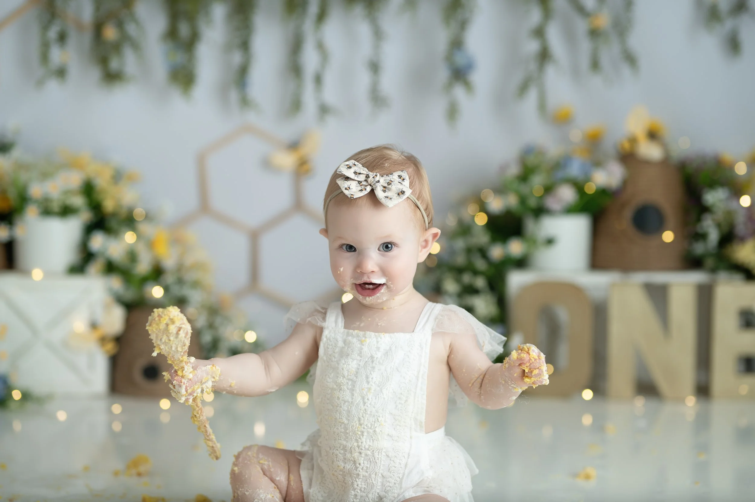 Baby girl smiling with cake-covered hands during a softly styled first birthday Cake Smash photography session.