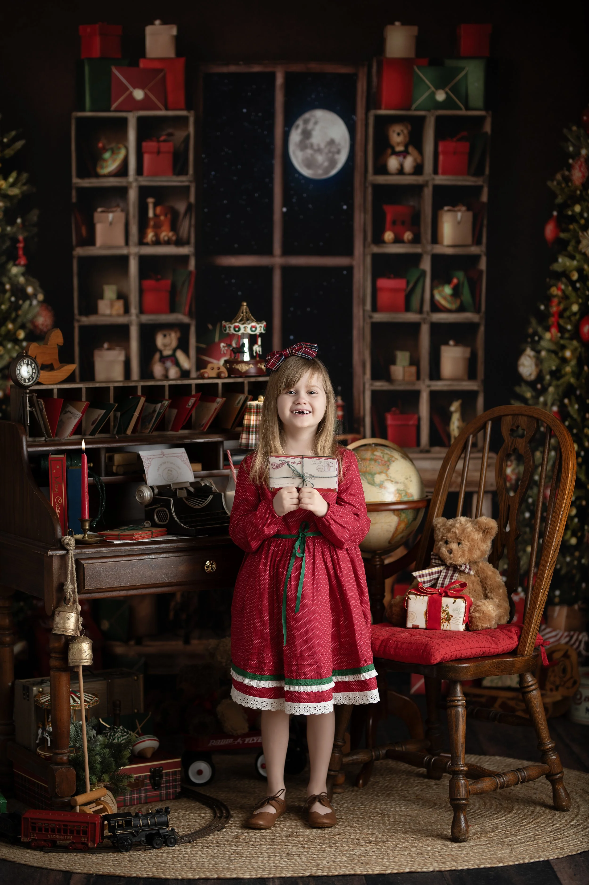 Young girl in a red holiday dress posing in a traditional Christmas photography set.