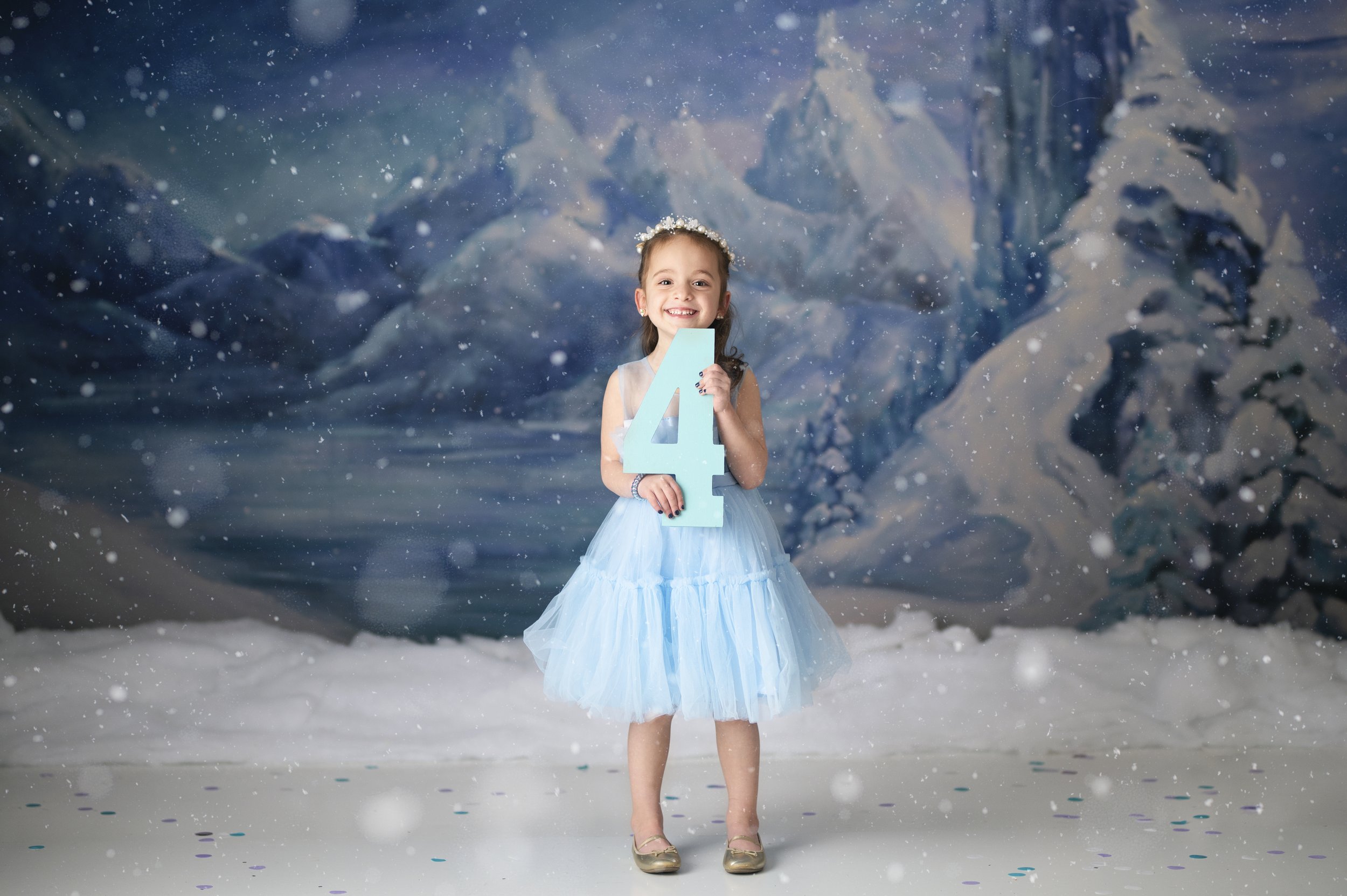 Young girl in a blue dress posing in a snowy winter-themed milestone photography set.