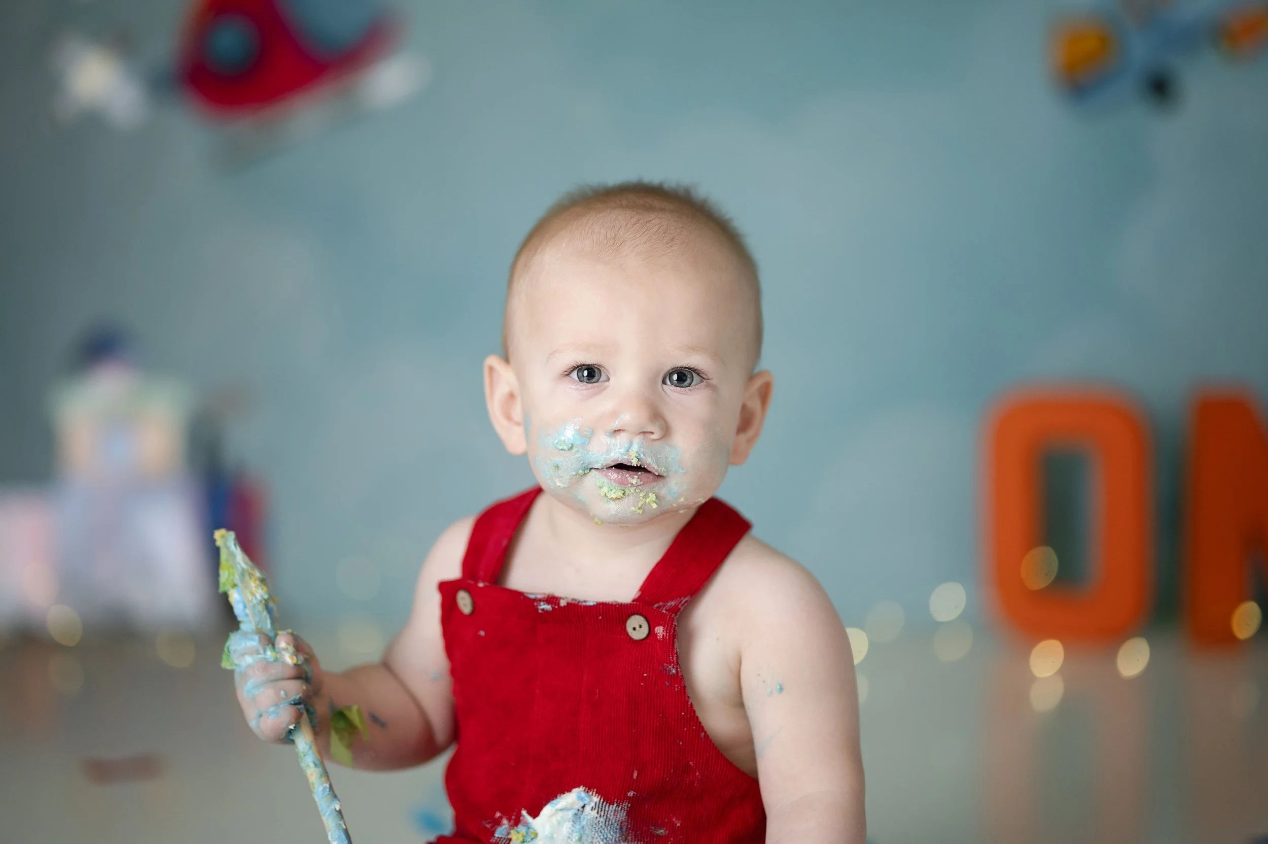 Baby in red overalls enjoying cake during a colorful car-themed birthday photography session.