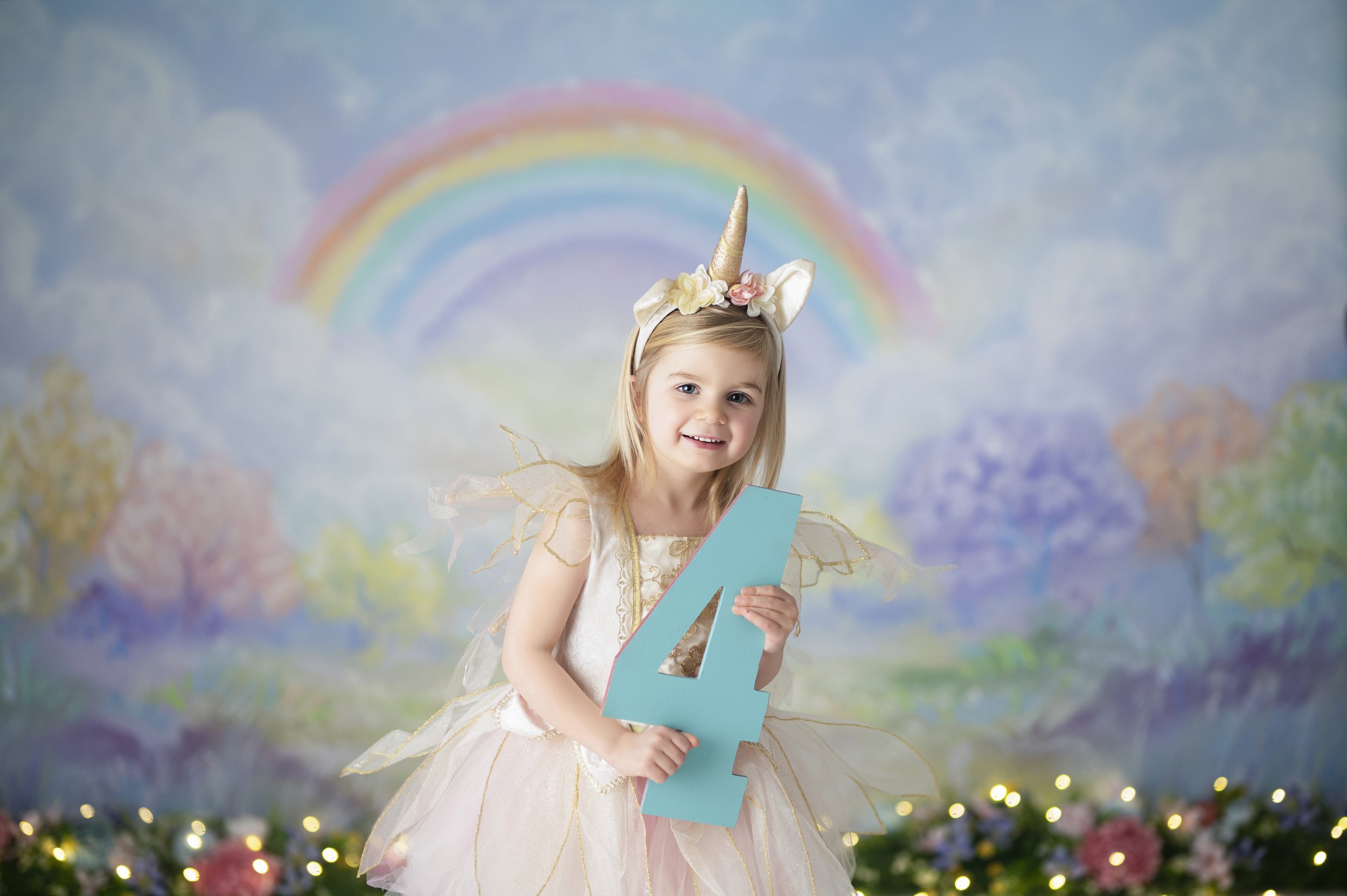 Young girl wearing a unicorn headband and holding a number four in front of a pastel rainbow backdrop.