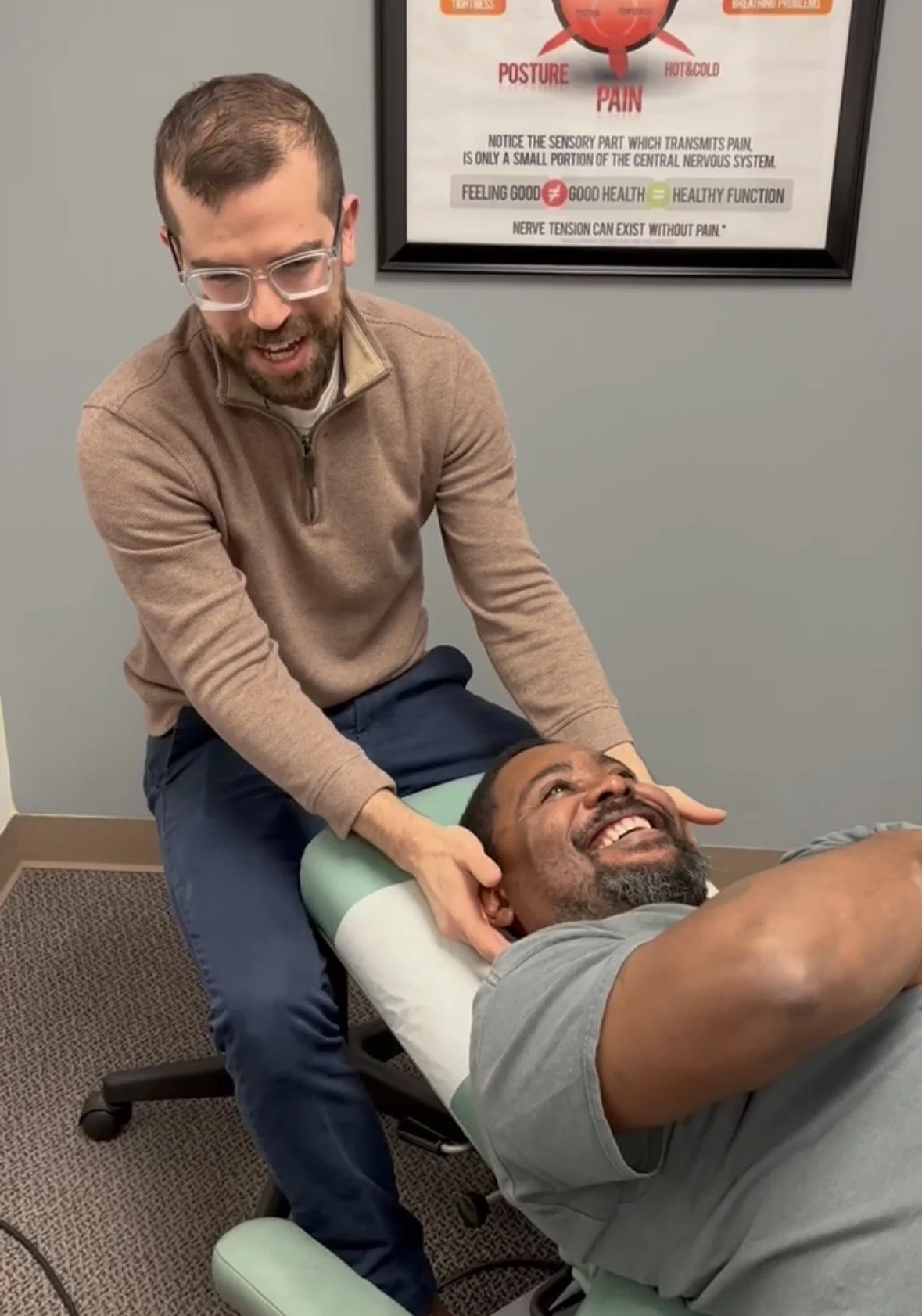 A chiropractor adjusts a patient's neck while the patient lies on a chiropractic table, both smiling in a clinic with a poster on back explaining pain and posture.