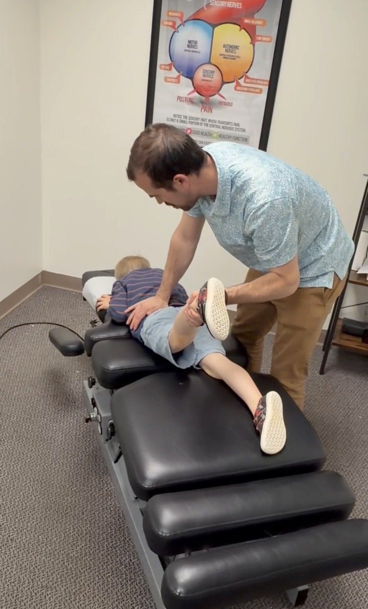 A chiropractor adjusting a young boy lying face down on a chiropractic table in a clinic. There is a poster about nerves on the wall behind them.