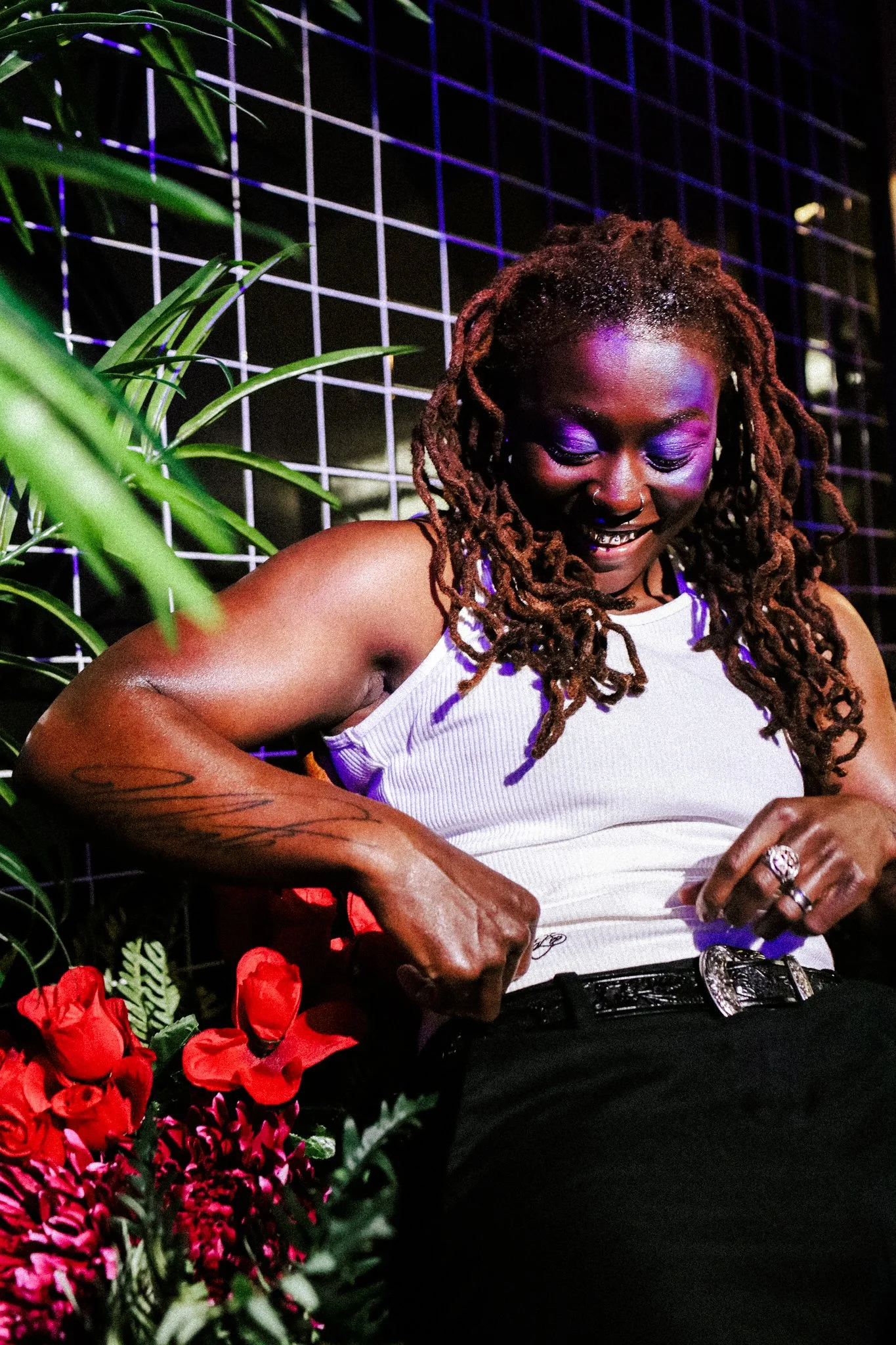 A smiling woman with dreadlocks and a nose piercing, wearing a white top and black pants with a belt, is sitting near plants with red flowers. She has a tattoo on her upper arm and rings on her fingers. The background features a black grid with purple lighting.