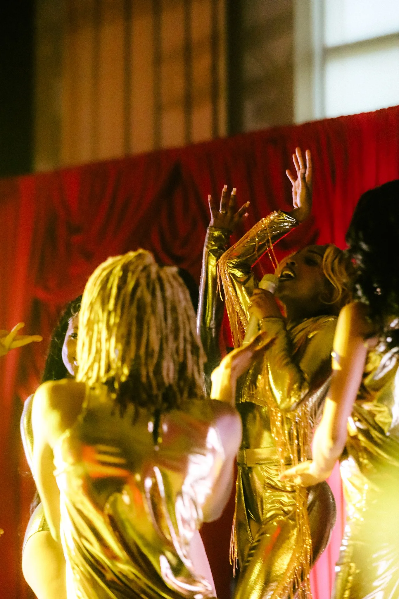 A group of people singing and dancing on a stage with red curtains, wearing shiny gold outfits, and raising their hands in the air.