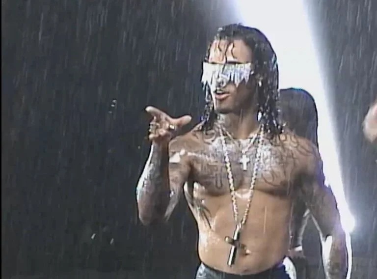 A shirtless man wearing sunglasses with fringes, necklaces, and tattoos, standing in the rain, making a hand gesture.