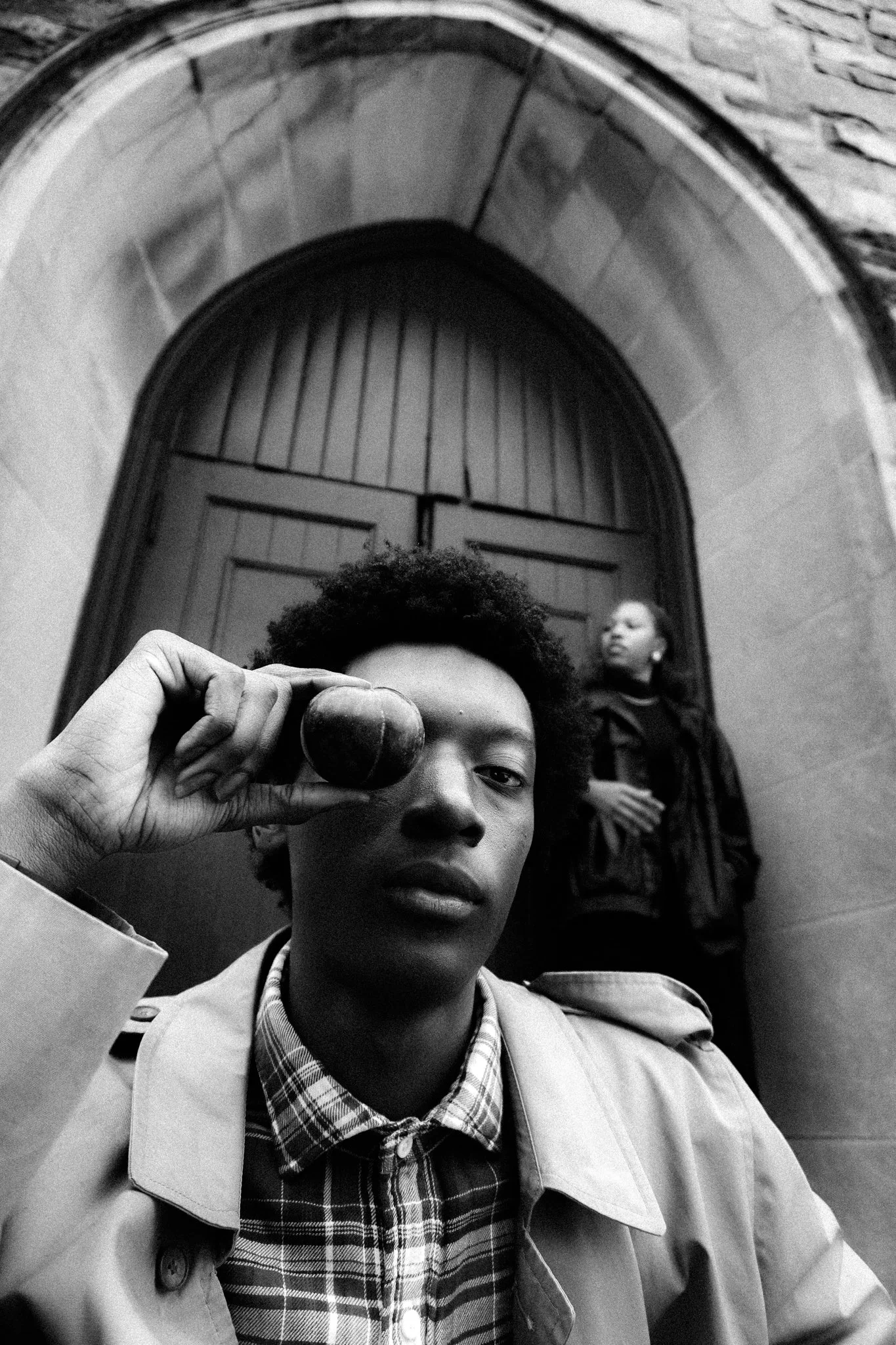 A young person with short curly hair holding a small round object up to their eye, while looking into a camera. In the background, a woman is standing near a wooden door in an arched stone entryway.