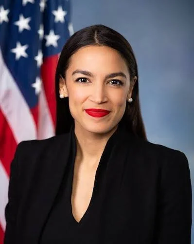 A woman with dark hair wearing a black blazer and red lipstick, standing in front of an American flag.