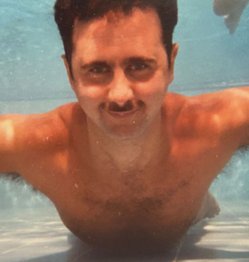 A man with dark hair and a mustache swimming in a pool, looking at the camera.