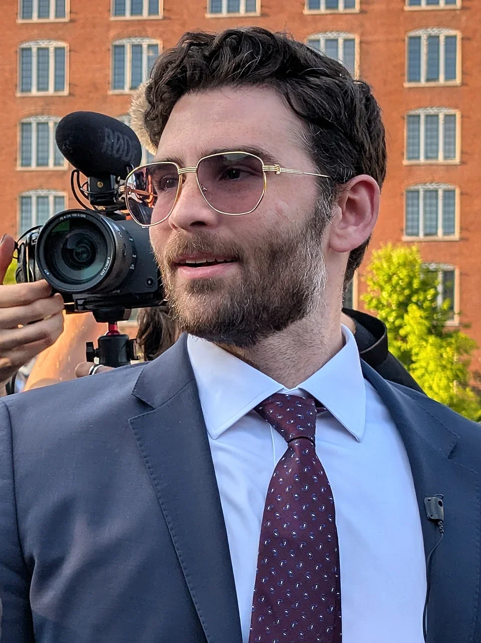A man in a suit and tie, wearing glasses, is standing outdoors with a camera crew behind him. There is a large brick building and some trees in the background.