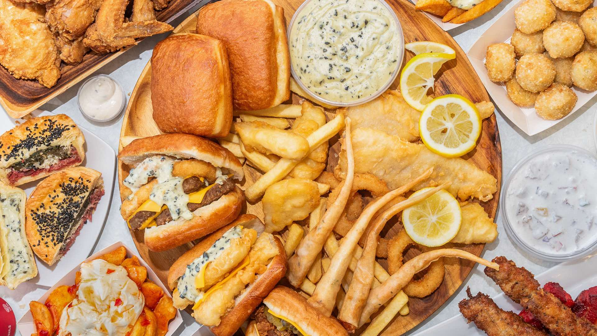 Variety of fried foods, sandwiches, potato fries, lemon slices, and dipping sauces on wooden and paper trays.