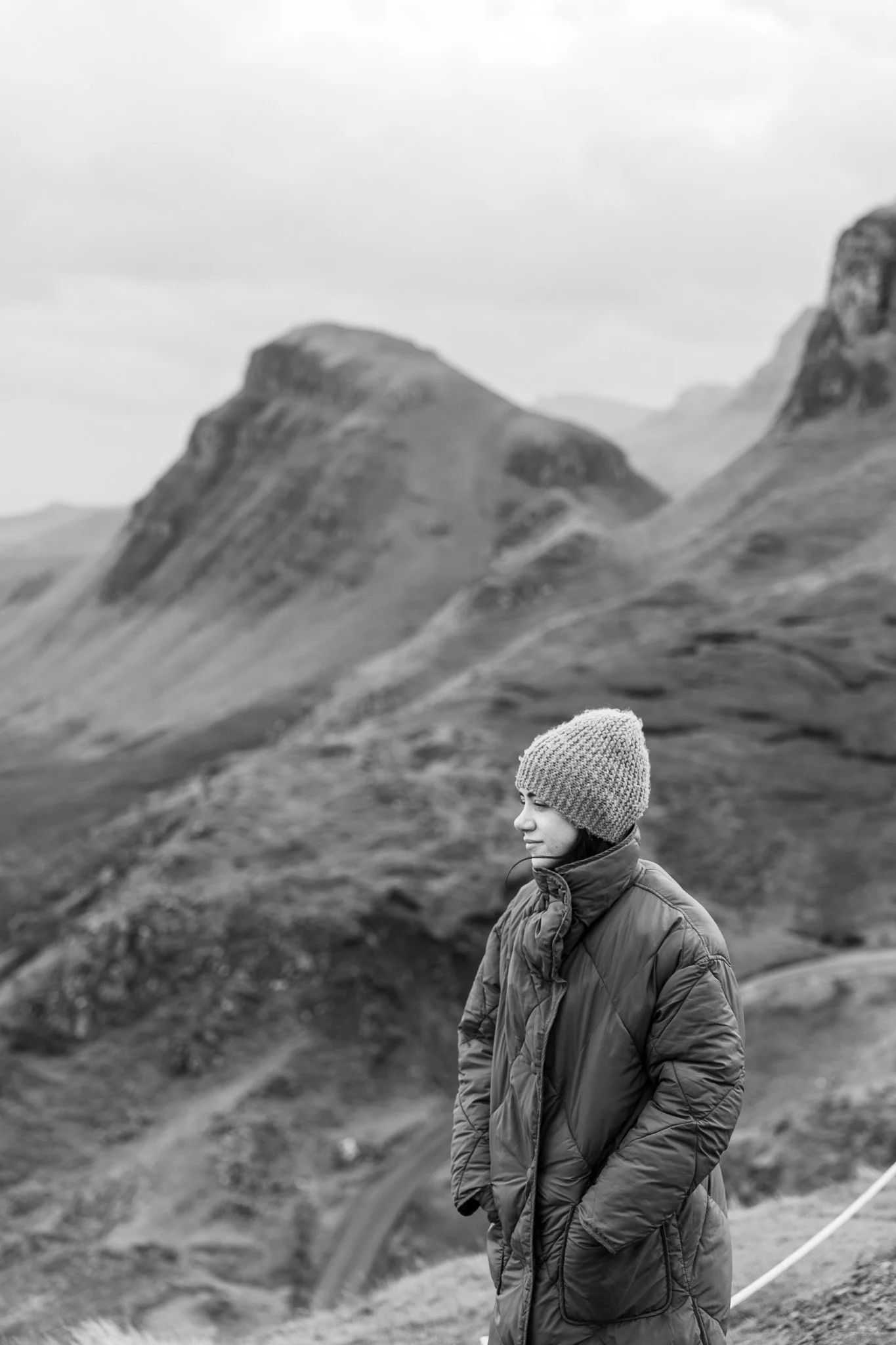 A woman wearing a knit hat and puffy jacket standing outdoors in a mountainous landscape with grassy hills and rocky peaks in black and white.