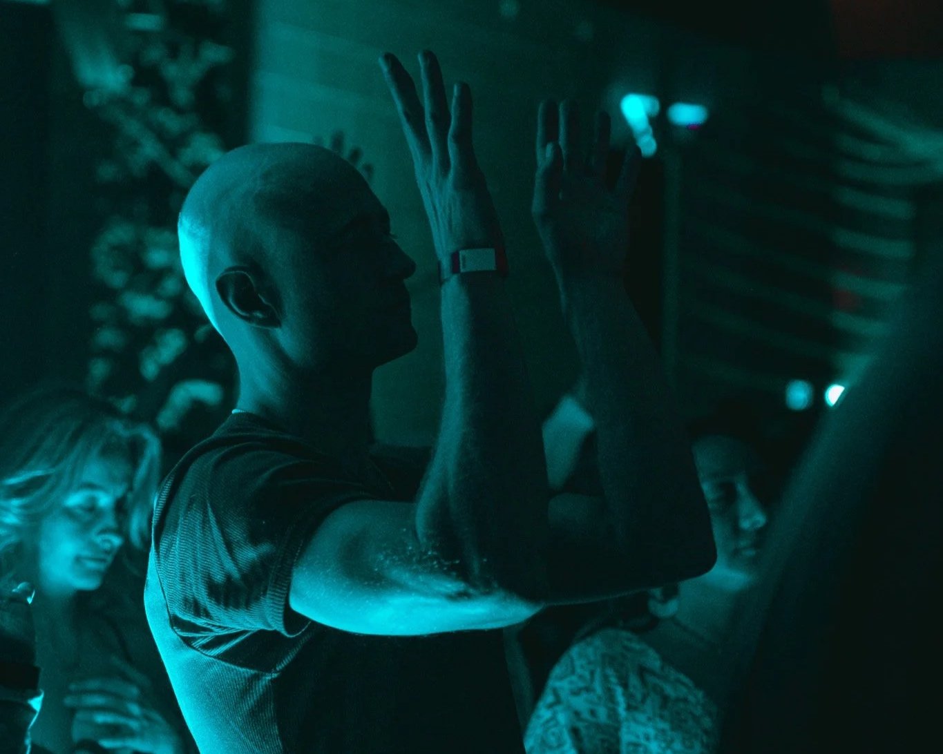 A group of people, including a bald man, standing in a dark room, with their hands pressed against a surface, illuminated by blue light.