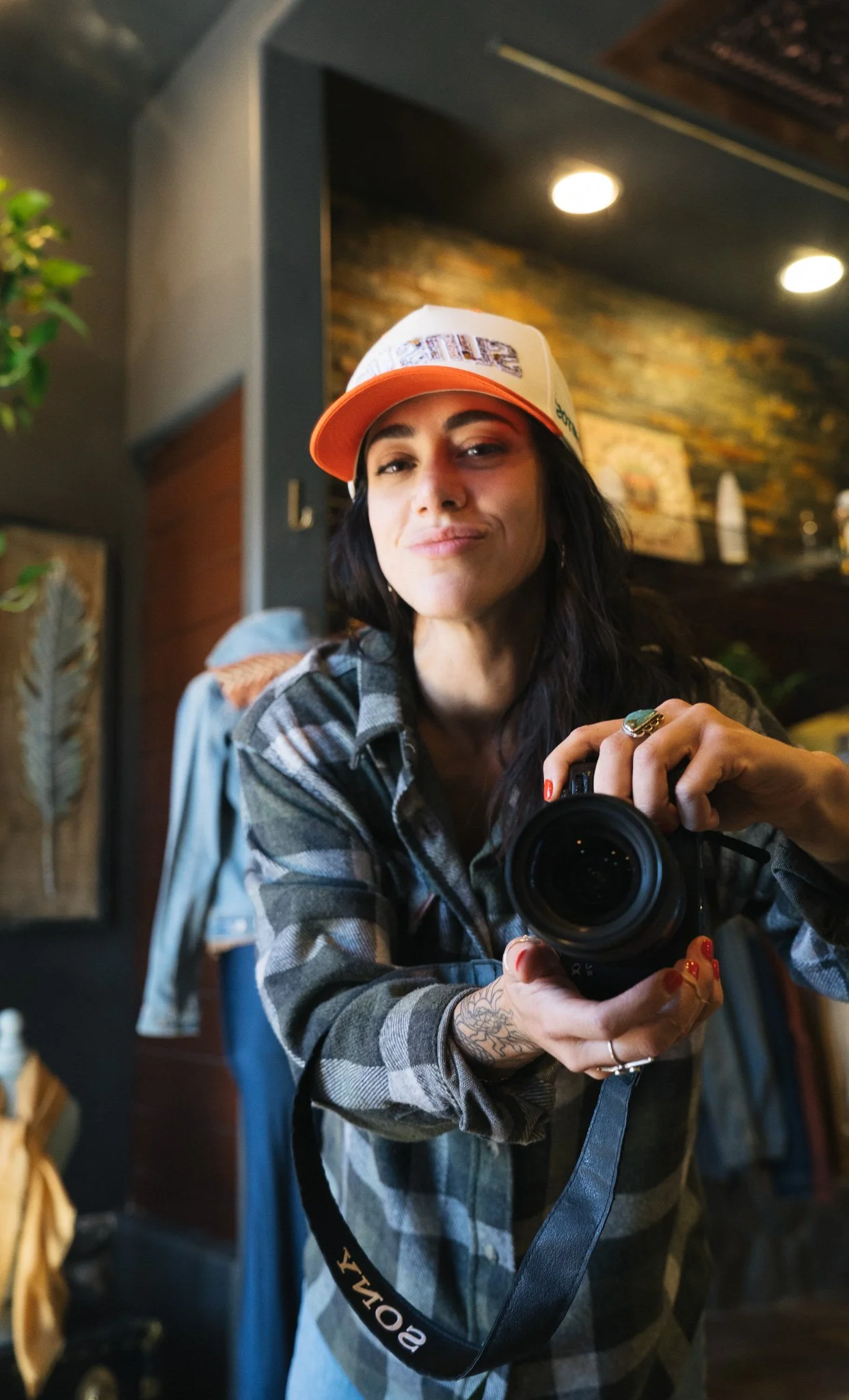 A woman taking a mirror selfie with a camera, wearing a trucker hat with the word 'PEACE' and a plaid shirt, in a dimly lit room with a brick wall background.