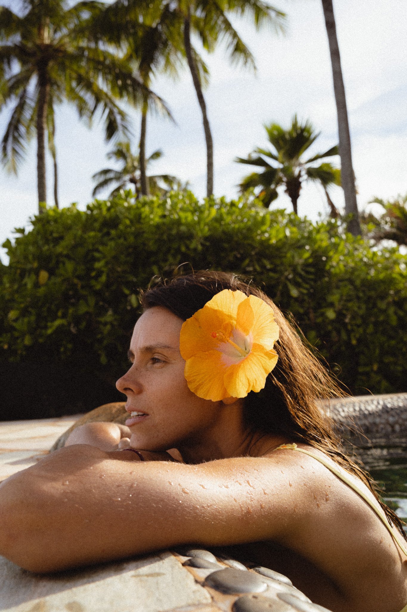 A woman with a yellow hibiscus flower in her hair, resting her arm on a stone surface, with palm trees and a cloudy sky in the background.