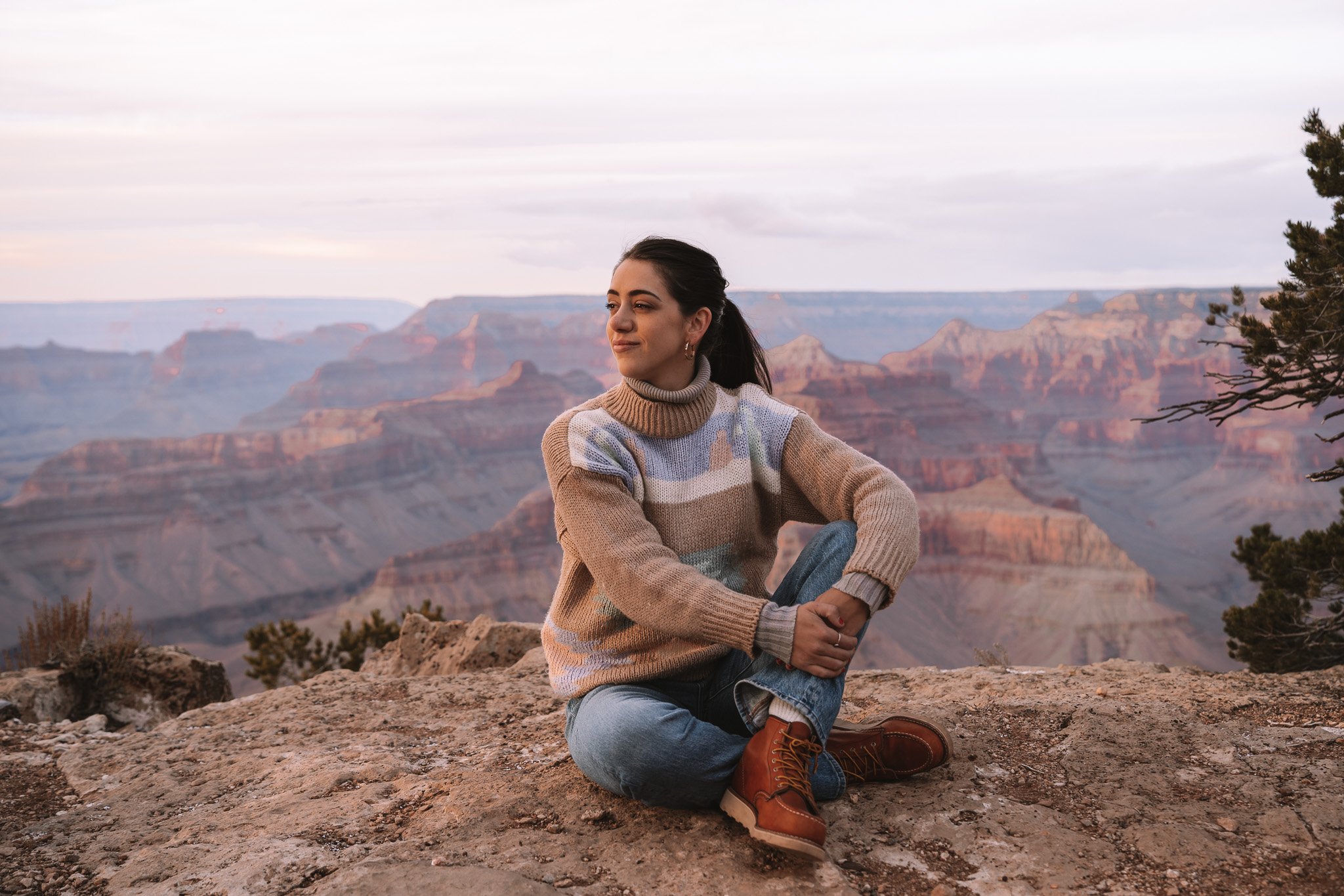 A woman sitting on a rocky ledge at the Grand Canyon during sunset, wearing a multicolored sweater, blue jeans, and brown boots.