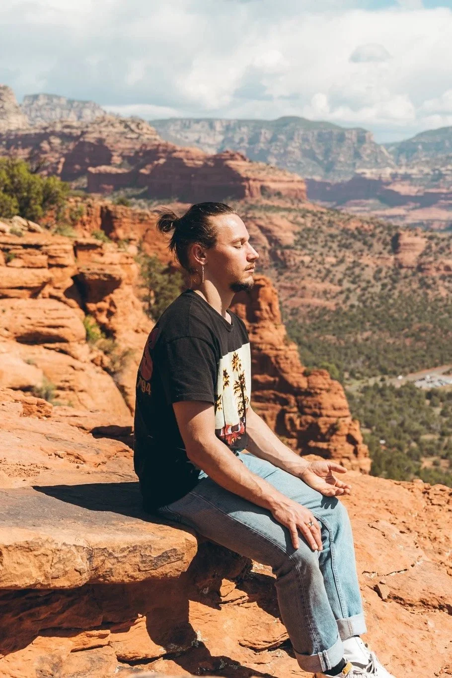 A man sitting on a rock ledge in a desert canyon landscape in Sedona, meditating with eyes closed, surrounded by red rock formations and distant mesas under a partly cloudy sky.