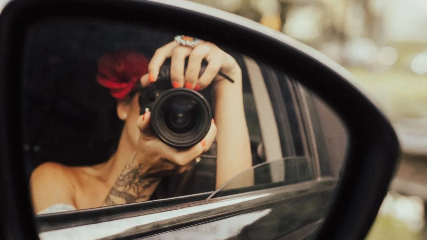 Person taking a photo with a camera in a car's side mirror, with a red flower in their hair and tattoos on their arm.