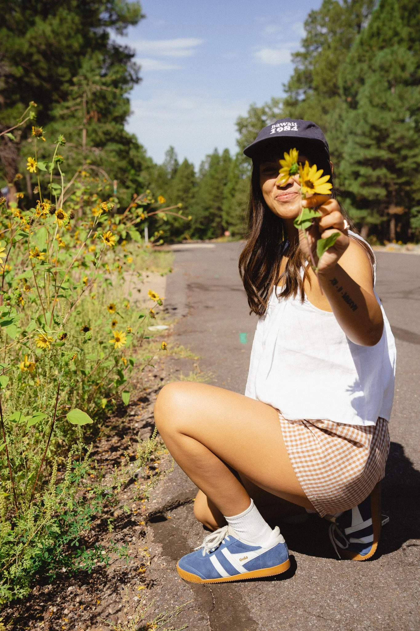 A young woman crouches on a sidewalk in a natural setting, holding a yellow flower in front of her face, wearing a baseball cap, a sleeveless top, checkered shorts, and blue sneakers.