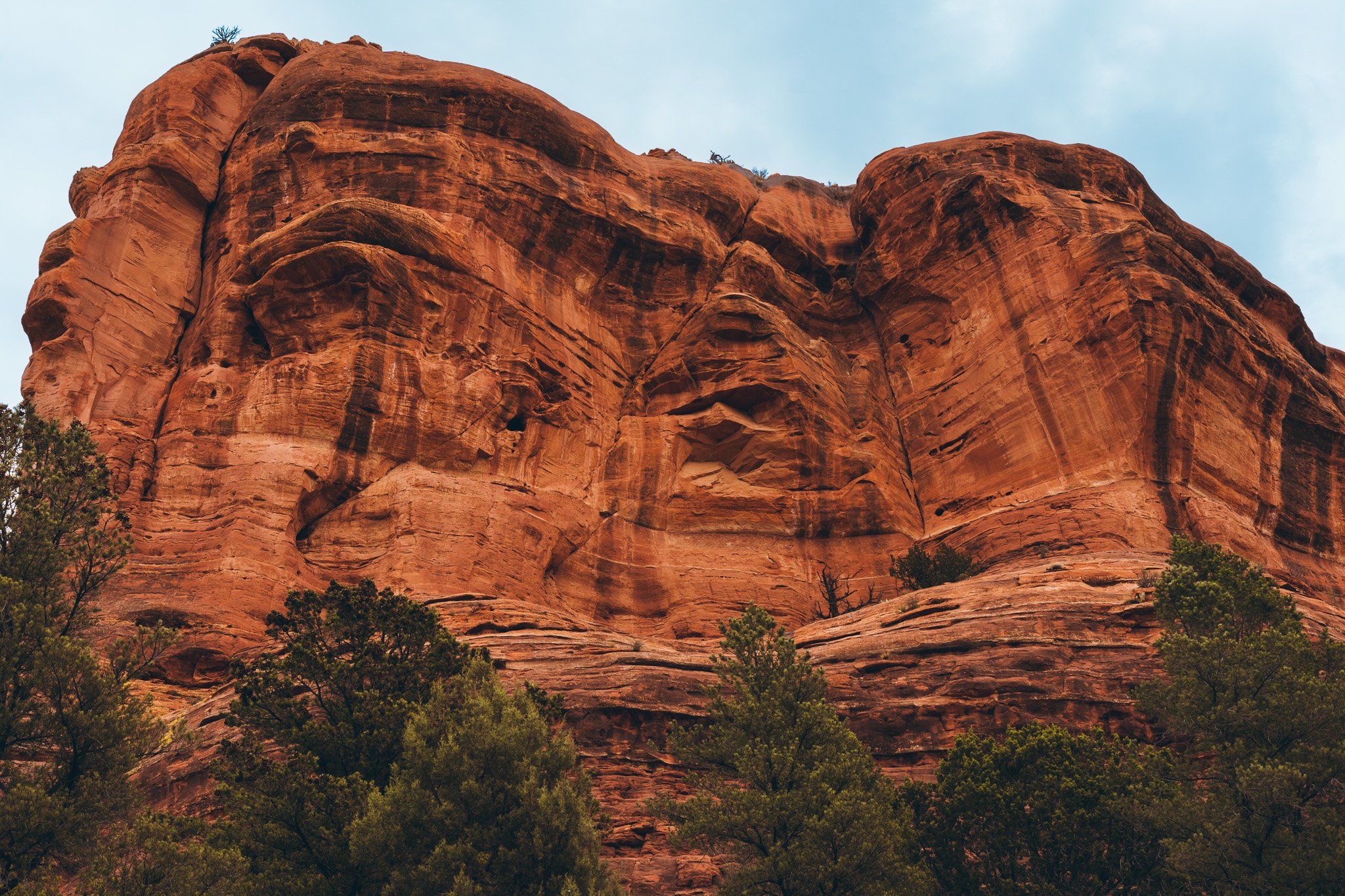 A large red sandstone rock formation with textured layers, with sparse trees and greenery at its base and a cloudy sky above.