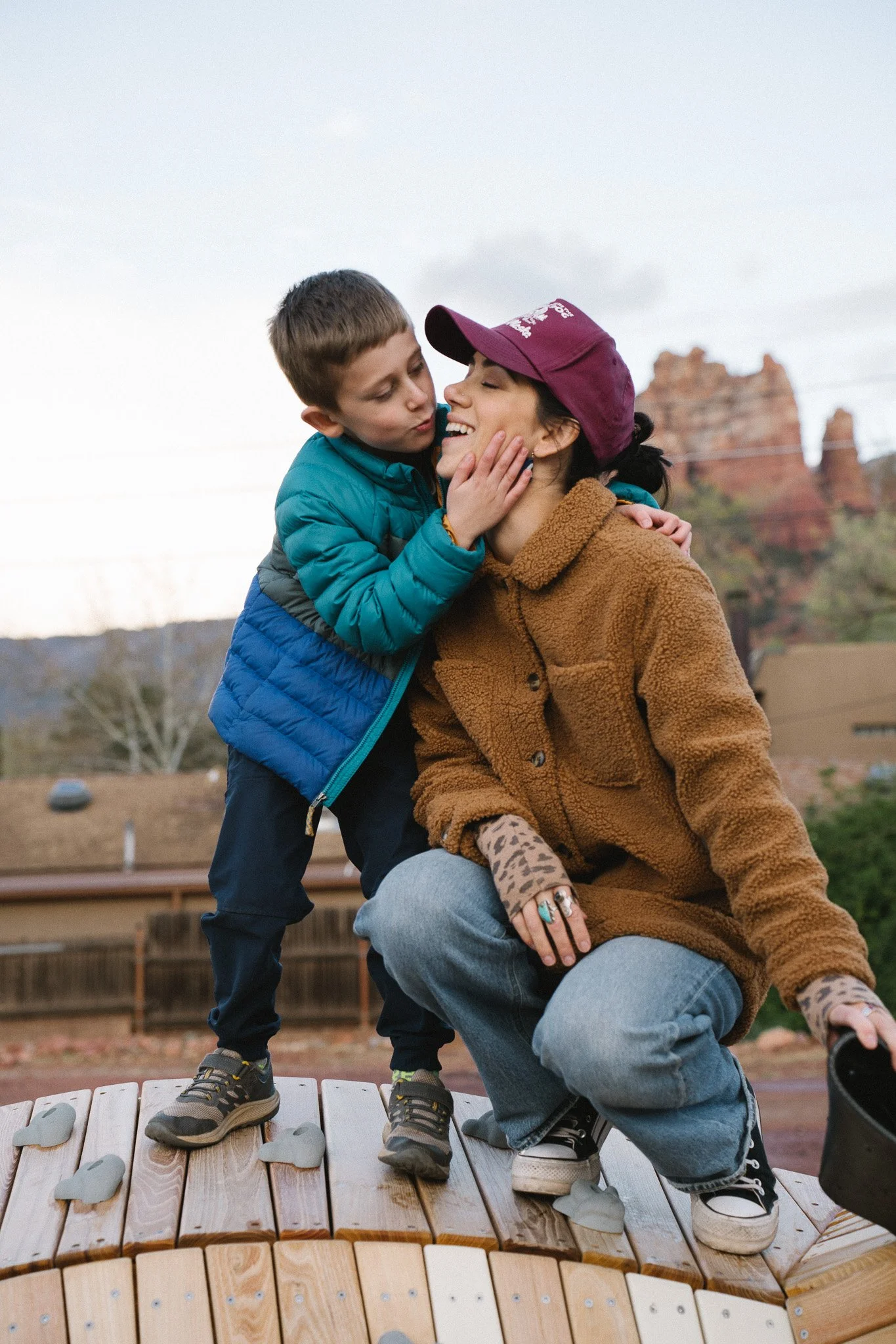 A woman and young boy share a joyful moment outdoors on a wooden platform with climbing rocks. The woman is kneeling, smiling, wearing a brown fleece jacket, gray pants, and a purple cap. The boy is standing close, wearing a blue puffer jacket and black shoes, touching the woman's face with one hand. The background shows a fence, trees, and a cloudy sky.