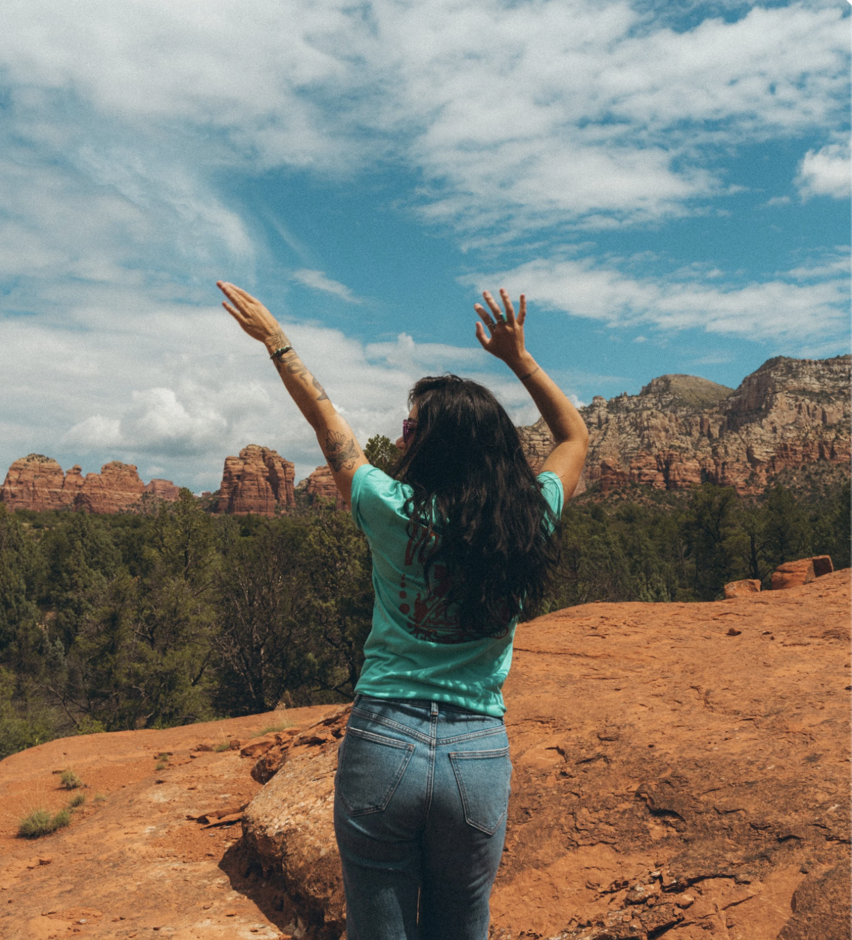 A woman with dark hair, tattoos, and sunglasses stands on a rocky landscape with red rock formations and green trees, raising her arms towards a blue sky with scattered clouds.