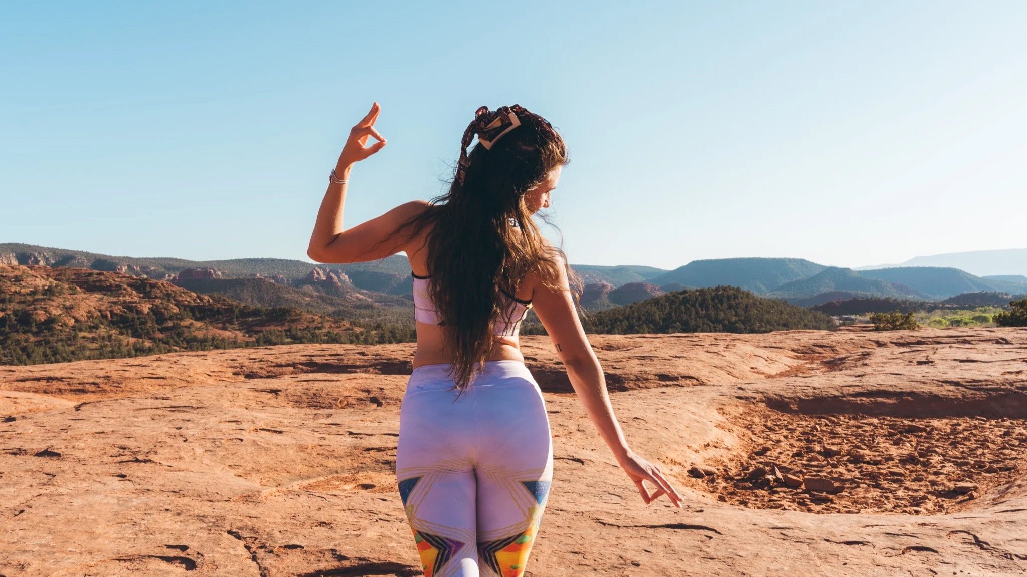 A woman practicing yoga outdoors on a rocky landscape with distant hills and clear sky, wearing a sports bra and colorful leggings.