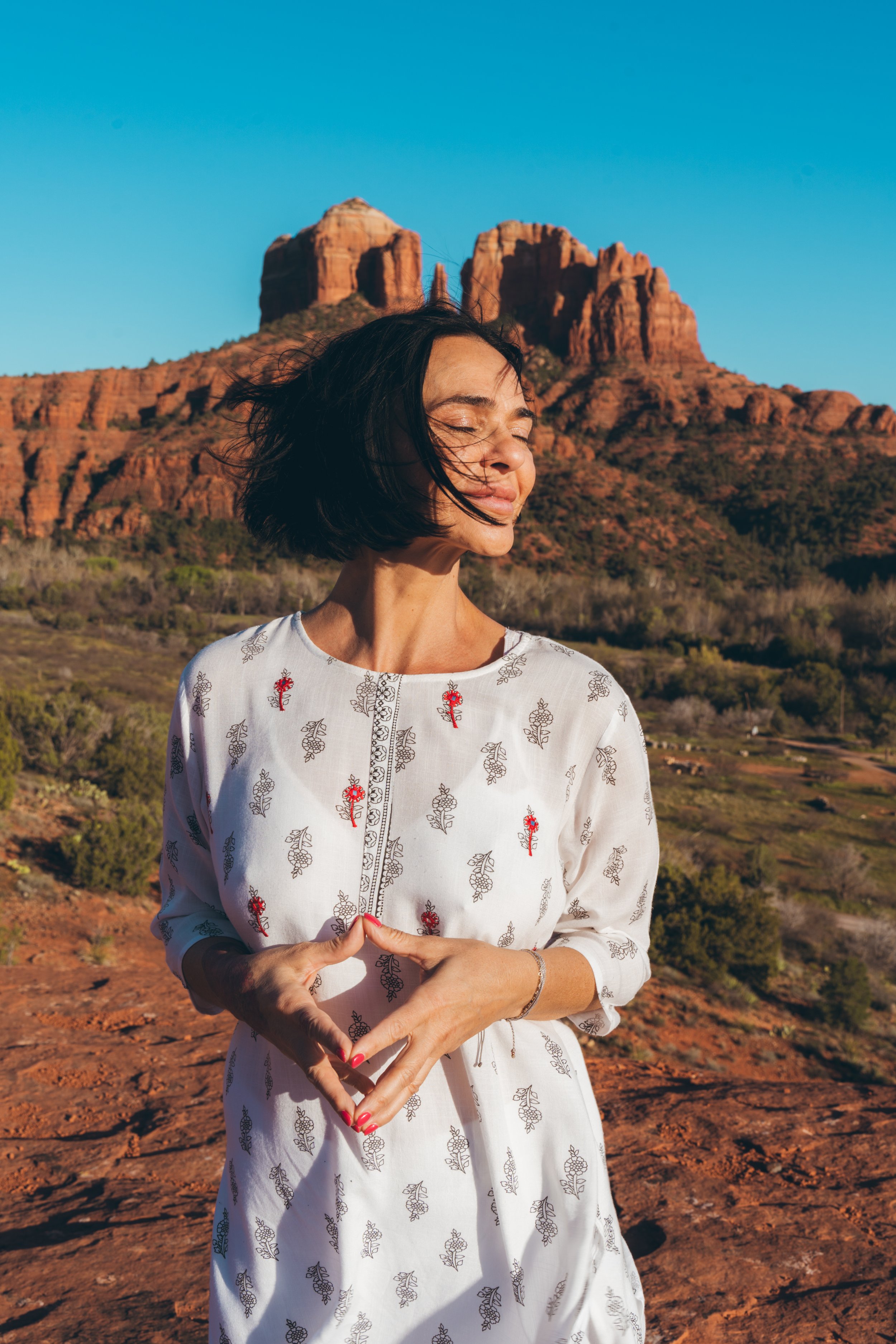 A woman with short dark hair, wearing a white dress with red and gray embroidery, stands outdoors with her eyes closed and a serene expression. She forms a heart shape with her hands in front of her. Behind her are red rock formations and a clear blu