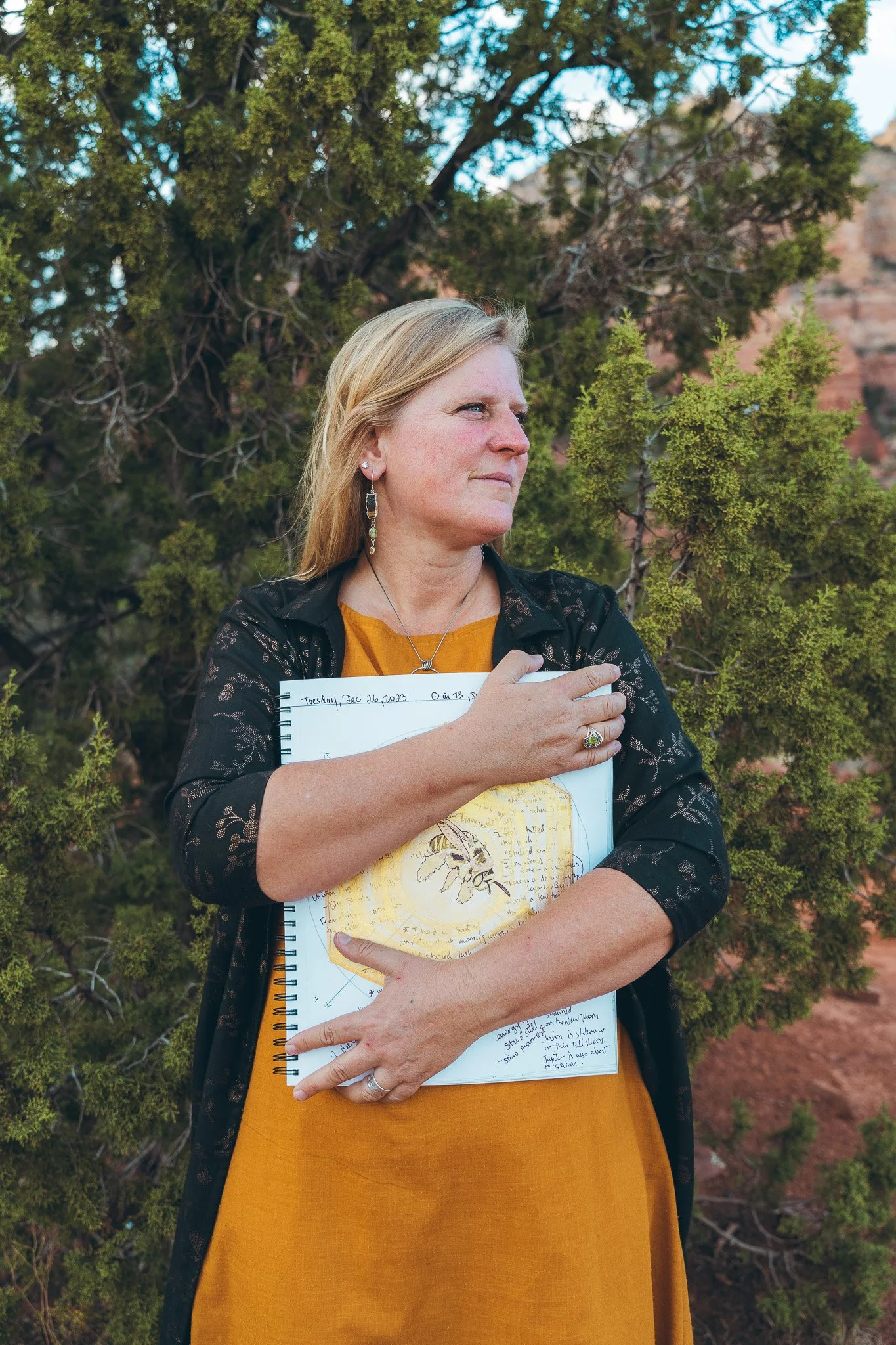 A woman with blonde hair holding a notebook with handwritten notes and a drawing of a bee, standing outdoors with trees and rocky formations in the background.