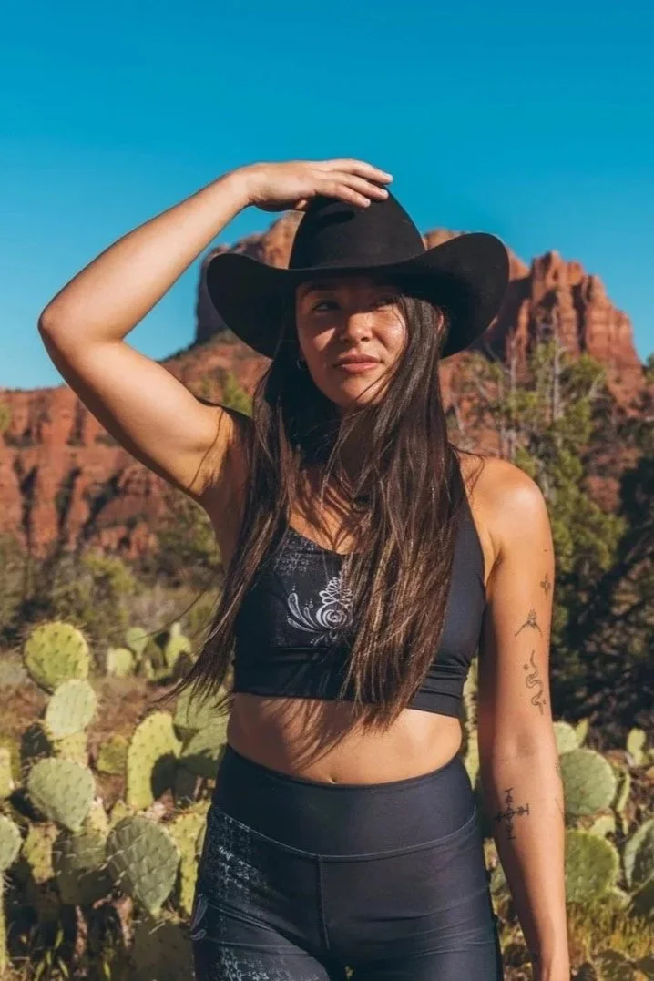 A woman in a cowboy hat standing outdoors in Sedona with cactus plants and red rock formations in the background.