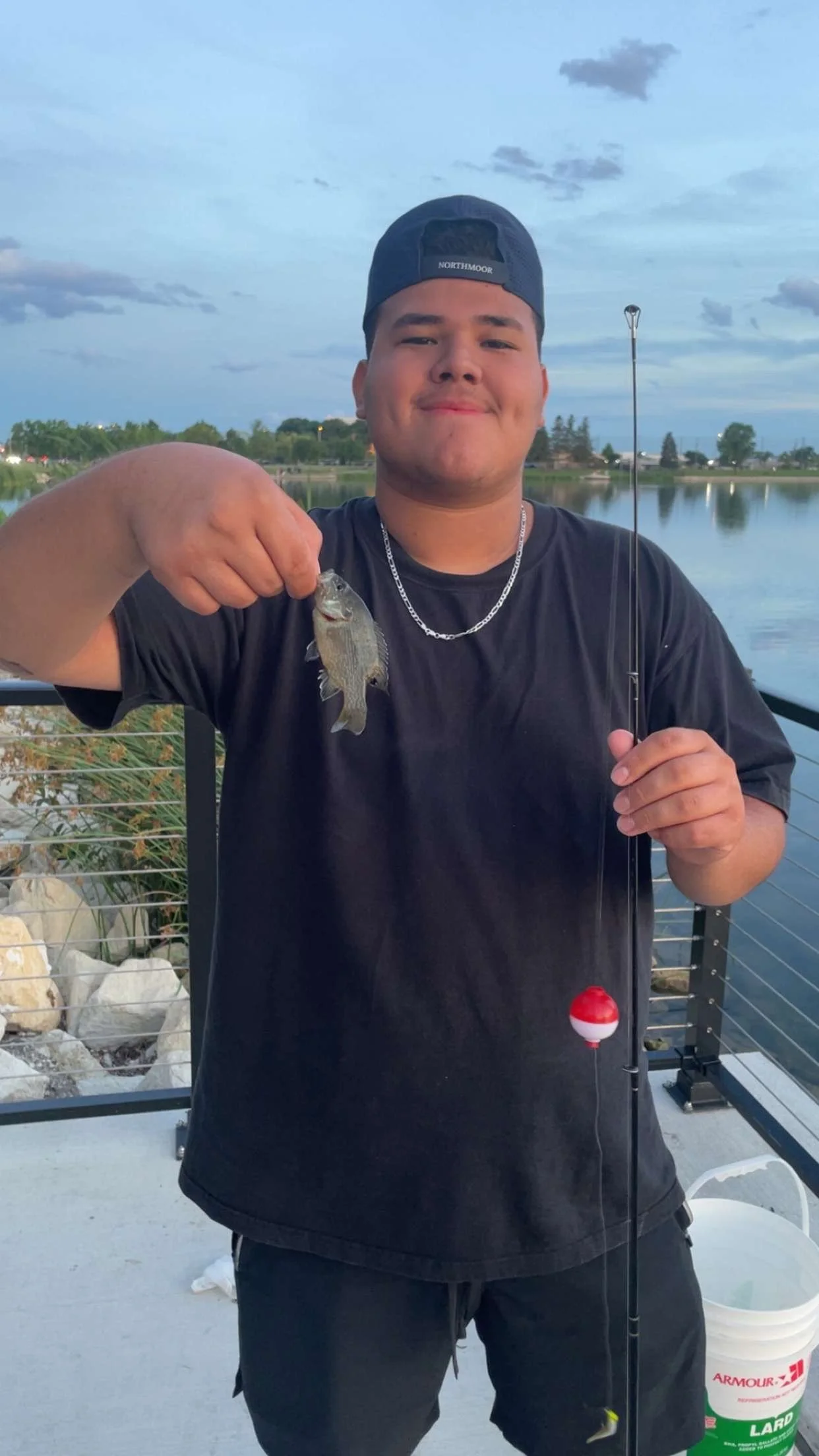 Young man wearing a black t-shirt and a backward cap, holding a small fish with one hand and a fishing rod with the other, standing near a body of water during evening.