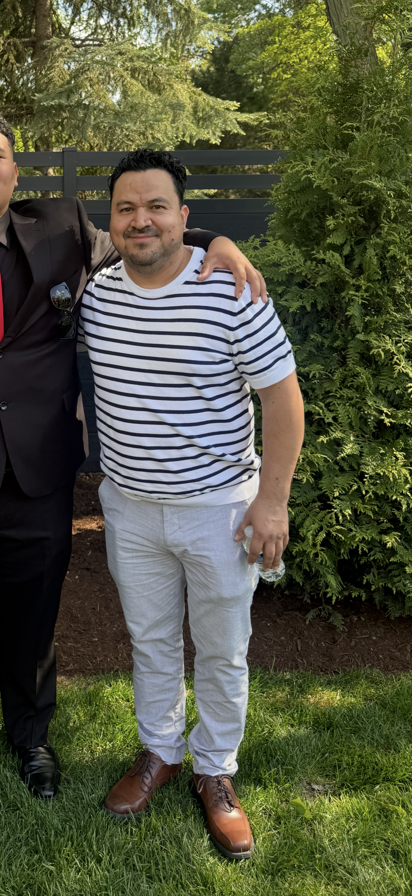 Man wearing a white and navy striped t-shirt, light-colored pants, and brown shoes, standing outdoors next to a bush. He is holding a water bottle in his right hand and smiling.