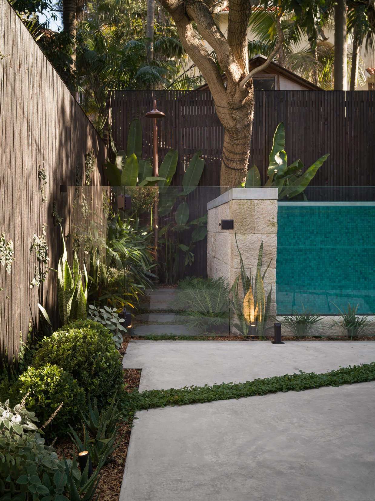 Luxury backyard landscape construction featuring a concrete swimming pool, glass pool wall, natural stone pillars, tropical planting and outdoor lighting