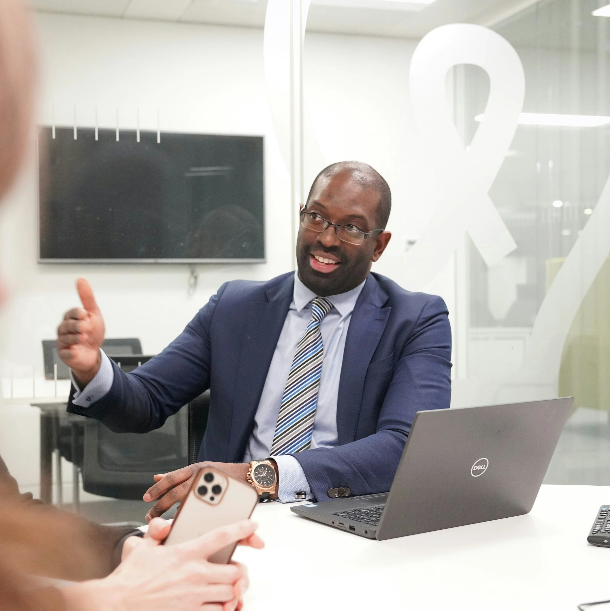 A man in a suit and striped tie is sitting at a desk with a Dell laptop open in front of him. Someone just handed him a phone, which he is holding in his left hand, and he is gesturing with his right thumb up. The setting appears to be a modern office with glass walls, and there is a TV monitor on the wall behind him.